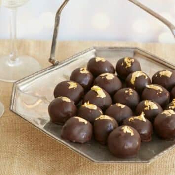 Heavenly Champagne Truffles are placed on a silver platter with a handle. they have bittersweet chocolate shells and are decorated with edible gold leaves. Two glasses of champagne are served on the side.