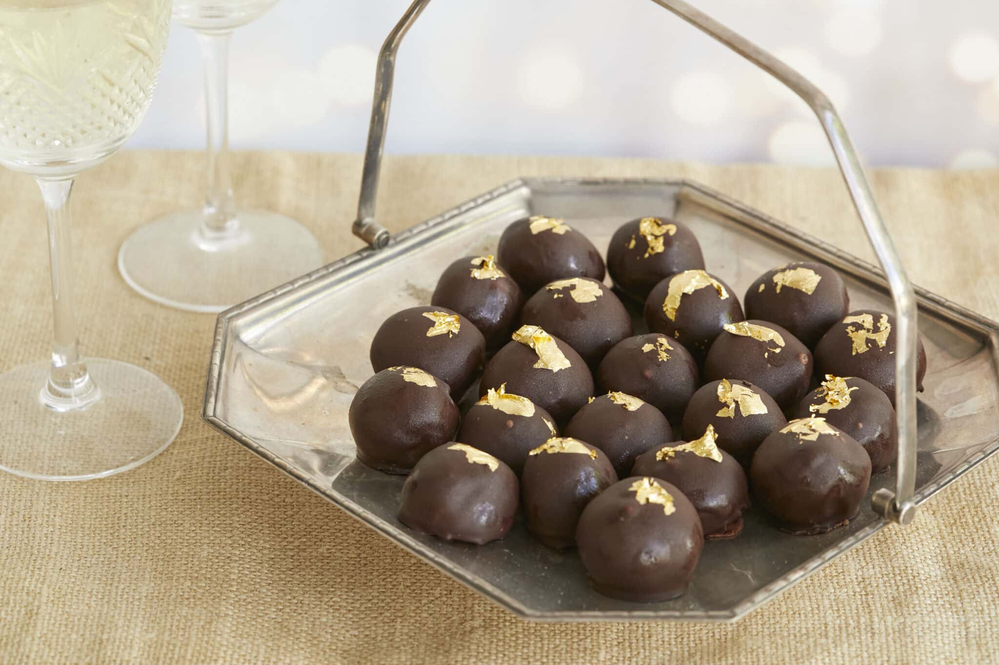 Heavenly Champagne Truffles are placed on a silver platter with a handle. they have bittersweet chocolate shells and are decorated with edible gold leaves. Two glasses of champagne are served on the side.
