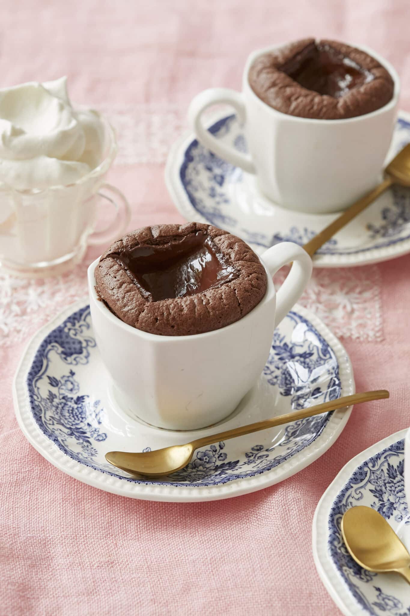 Baked hot chocolate is served in two white mugs on blue floral saucers with golden spoons. A glass cup of whipped cream is on the side. It's both light and rich, with an addictive contrast between the melty center and the surrounding soufflé-like cake.