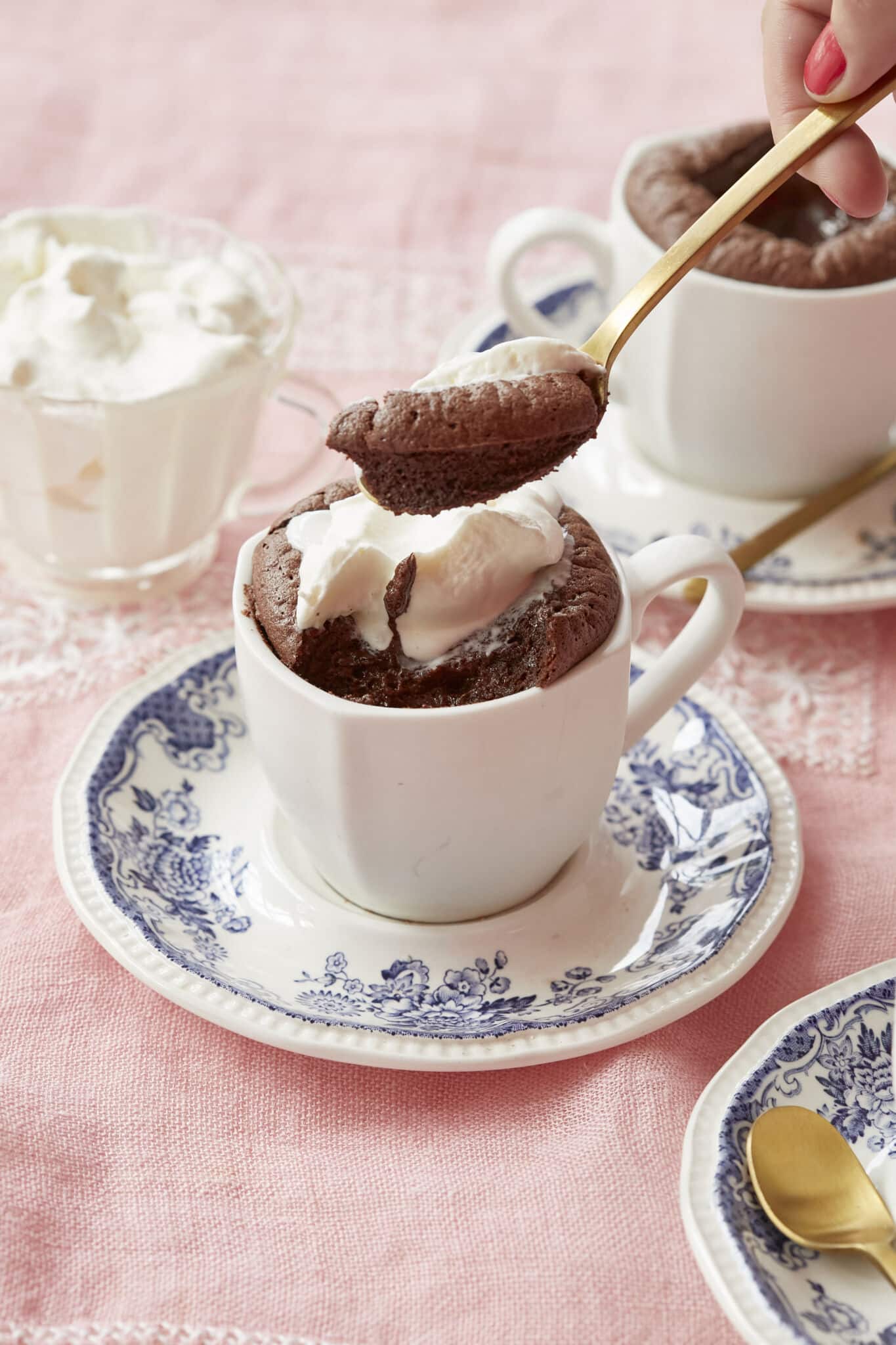 Baked hot chocolate is served in two white mugs on blue floral saucers with golden spoons. A glass cup of whipped cream is on the side. It's both light and rich, with an addictive contrast between the melty center and the surrounding soufflé-like cake. One mug of the baked hot chocolate is topped with softly whipped cream. One spoonful of scooped baked chocolate shows the moist interior.