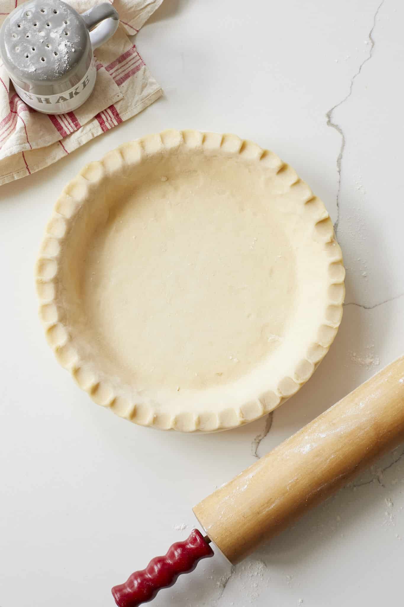 The Ballymaloe Creamed Butter Pie Crust is assembled into a pie pan with fluted edge. The flour shaker s in the top left corner, the wooden rolling pin with red handles is on the right side. 