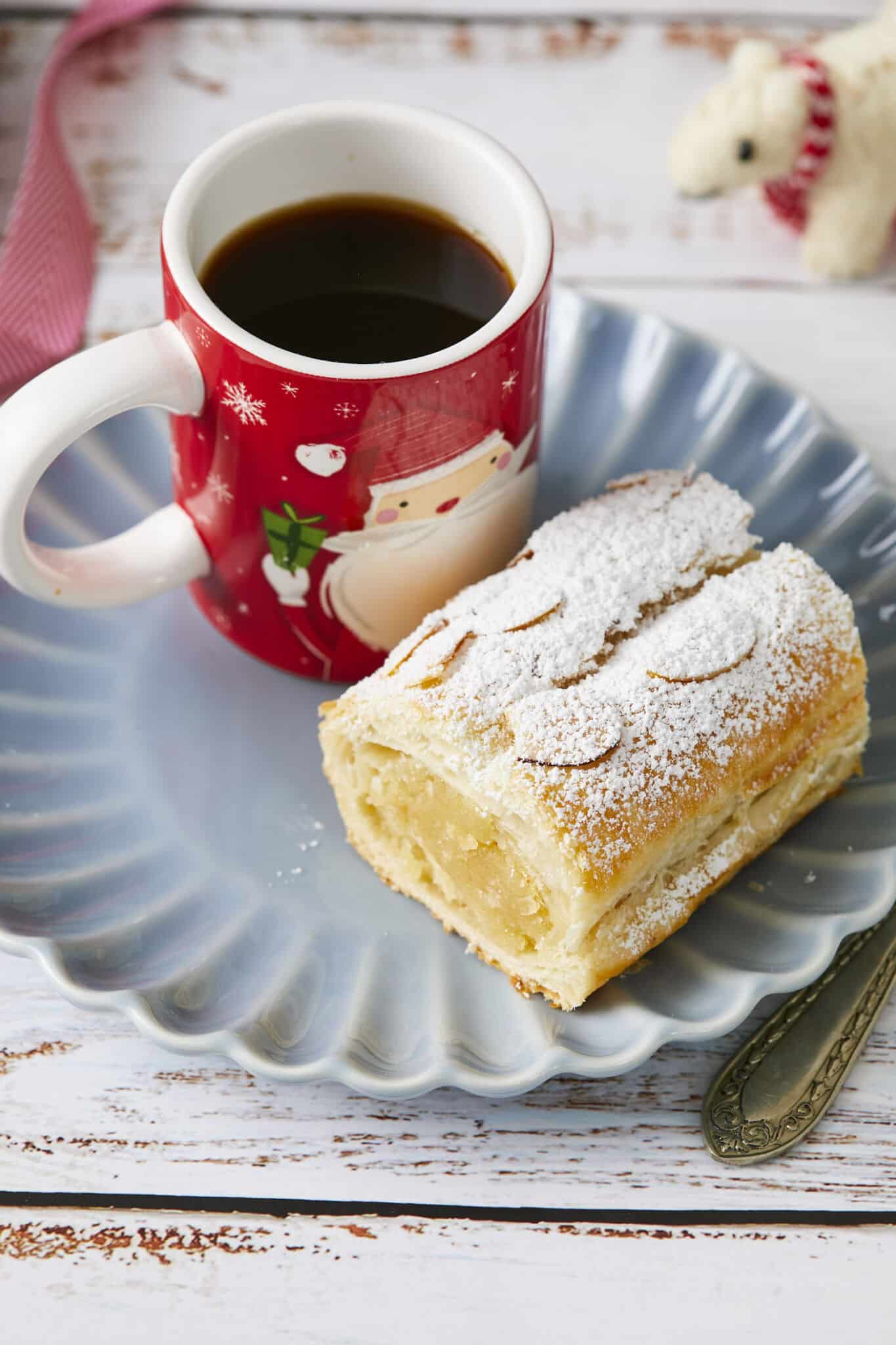 The delicate Dutch Banketstaaf is made from almond-paste-filled puff pastry logs topped with powdered sugar and sliced almonds and served sliced. It's served on a blue plate with a cup of coffee. 