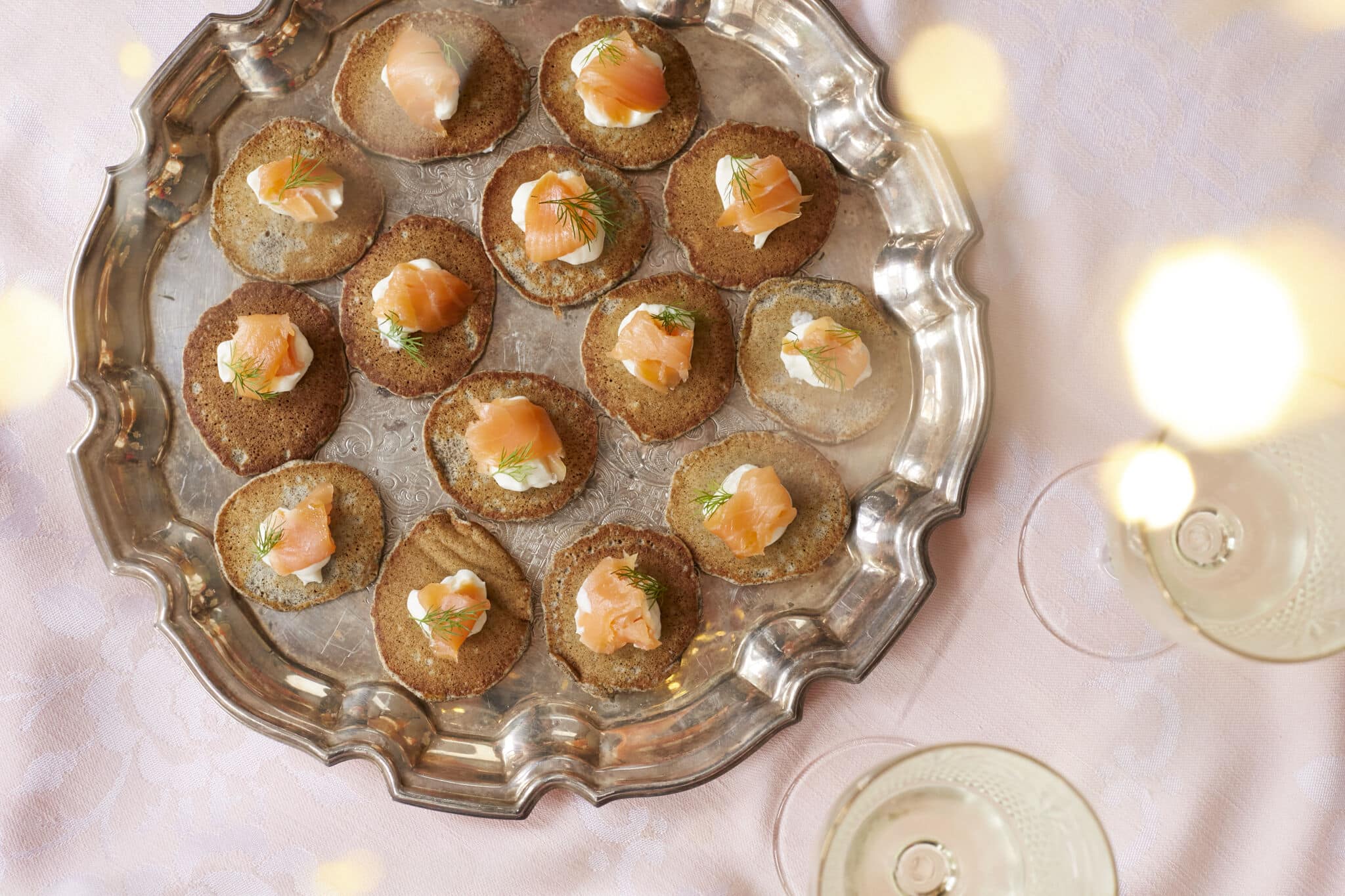 Small buckwheat pancakes, are fried and topped with crème fraîche and smoked salmon, served on a big silver platter.