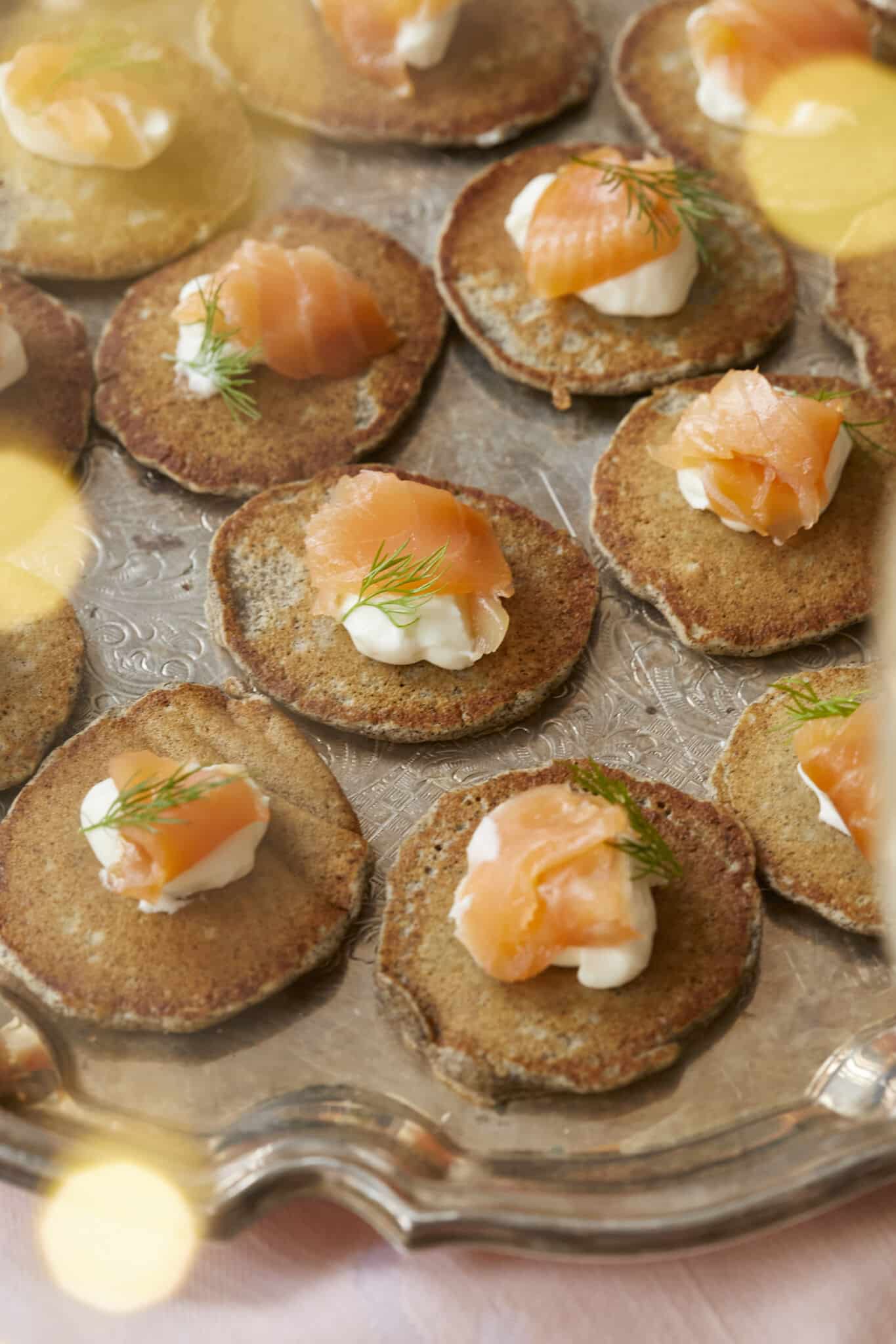 Small buckwheat pancakes, are fried and topped with crème fraîche and smoked salmon, served on a big silver platter. 