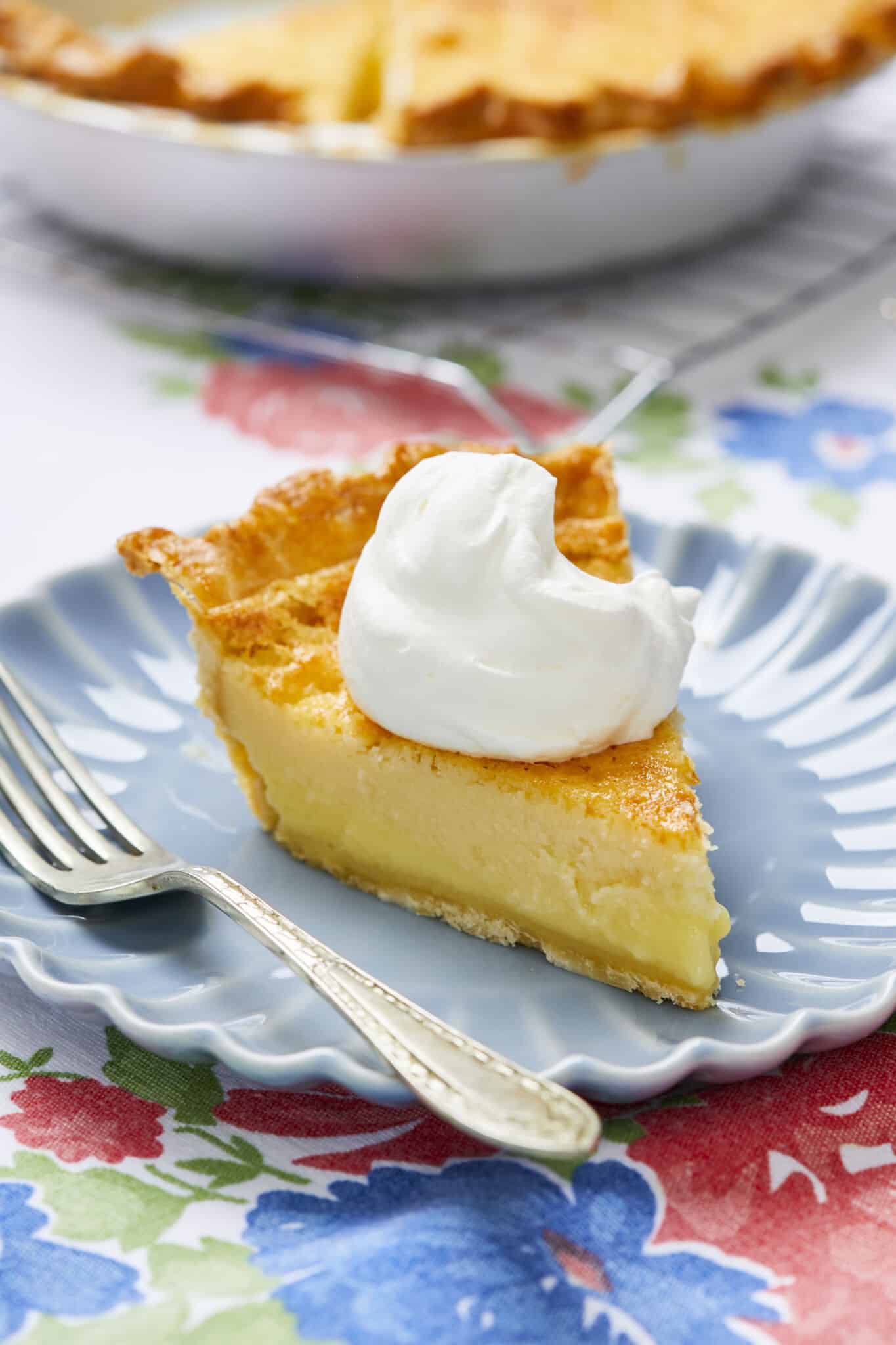The Irresistible Buttermilk Pie is baked in an aluminum pie dish. One slice is served on a blue dessert plate, with flaky crust, silky custard filling with caramelized top, crowned with freshly whipped cream.