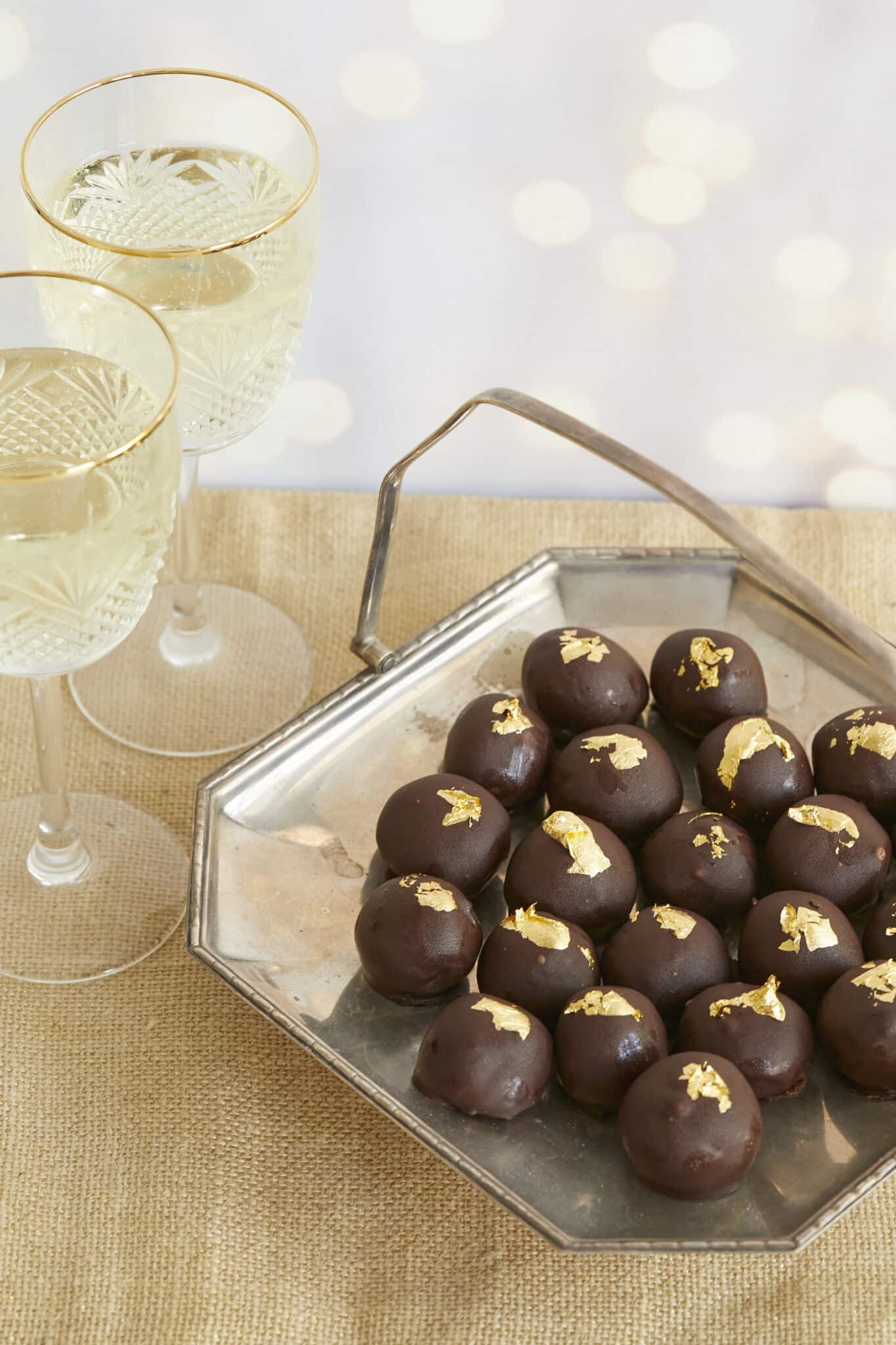 Heavenly Champagne Truffles are placed on a silver platter with a handle. they have bittersweet chocolate shells and are decorated with edible gold leaves. Two glasses of champagne are served on the side. 