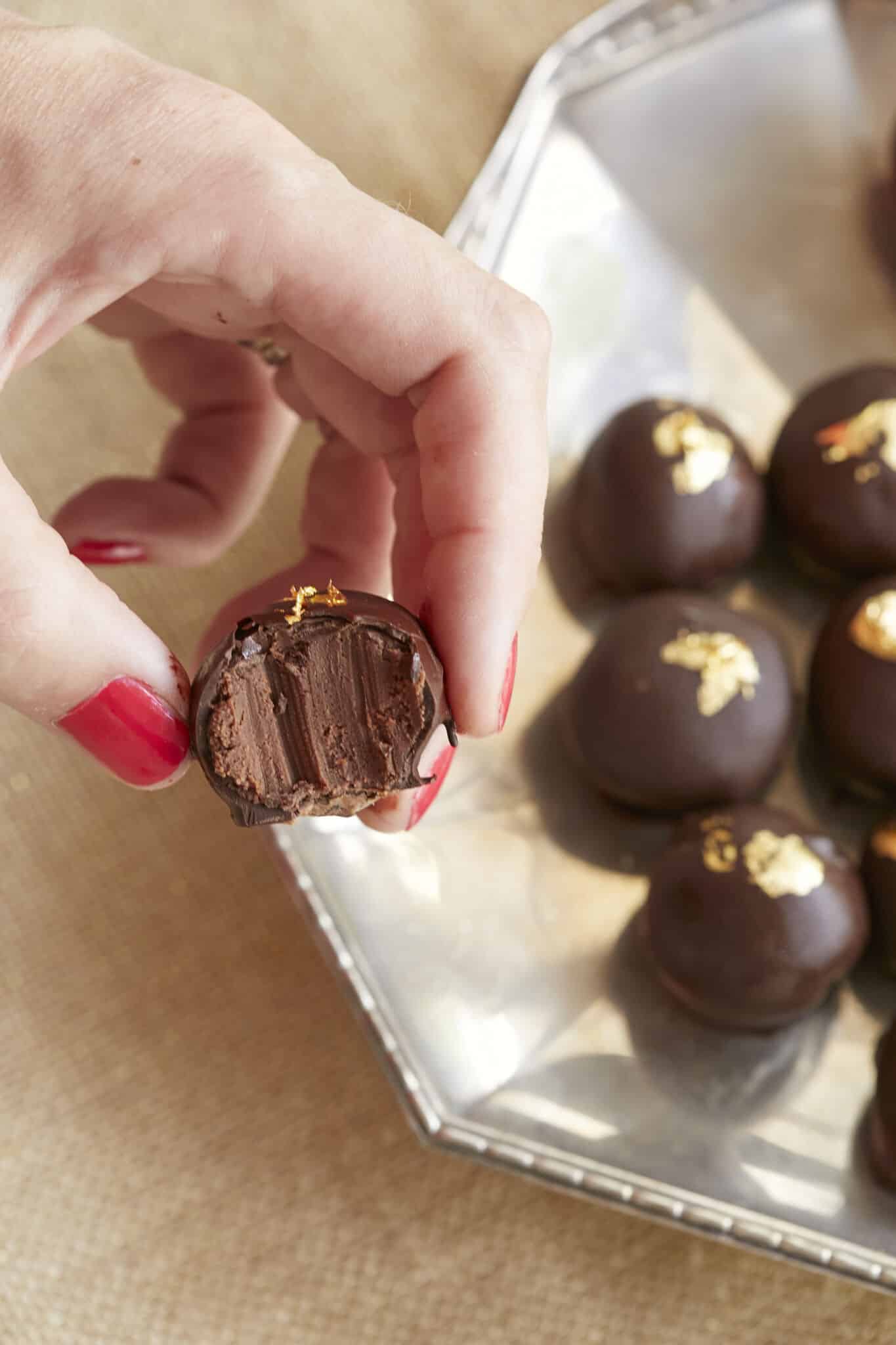 Heavenly Champagne Truffles are placed on a silver platter. They have bittersweet chocolate shells and are decorated with edible gold leaves. One truffle has been taken a bite and it shows the velvety smooth inside. 
