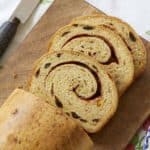 A loaf of Cinnamon Swirl Raisin Bread is sliced and served on a wooden board with butter on the side. The bread has a golden-brown and slightly crispy crust, a tender and even crumb, scattered raisins, and cinnamon swirls throughout upon slicing.