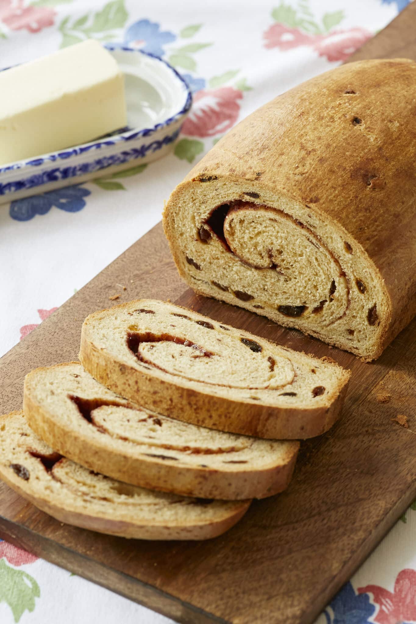 A loaf of Cinnamon Swirl Raisin Bread is sliced and served on a wooden board with butter on the side. The bread has a golden-brown and slightly crispy crust, a tender and even crumb, scattered raisins, and cinnamon swirls throughout upon slicing.