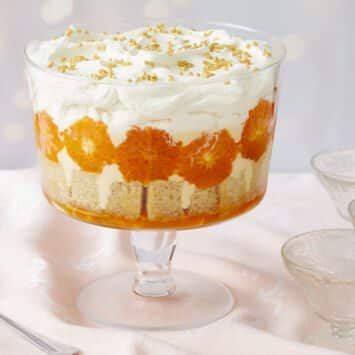A stunning Sweet and Zesty Clementine Trifle with Boozy Amaretto consists of layers of clementine syrup, cubed sponge cake, caramelized clementines, crème anglaise, lemon curd, and Amaretto whipped cream. Three glass dessert cups and one serving spoon are on the side.