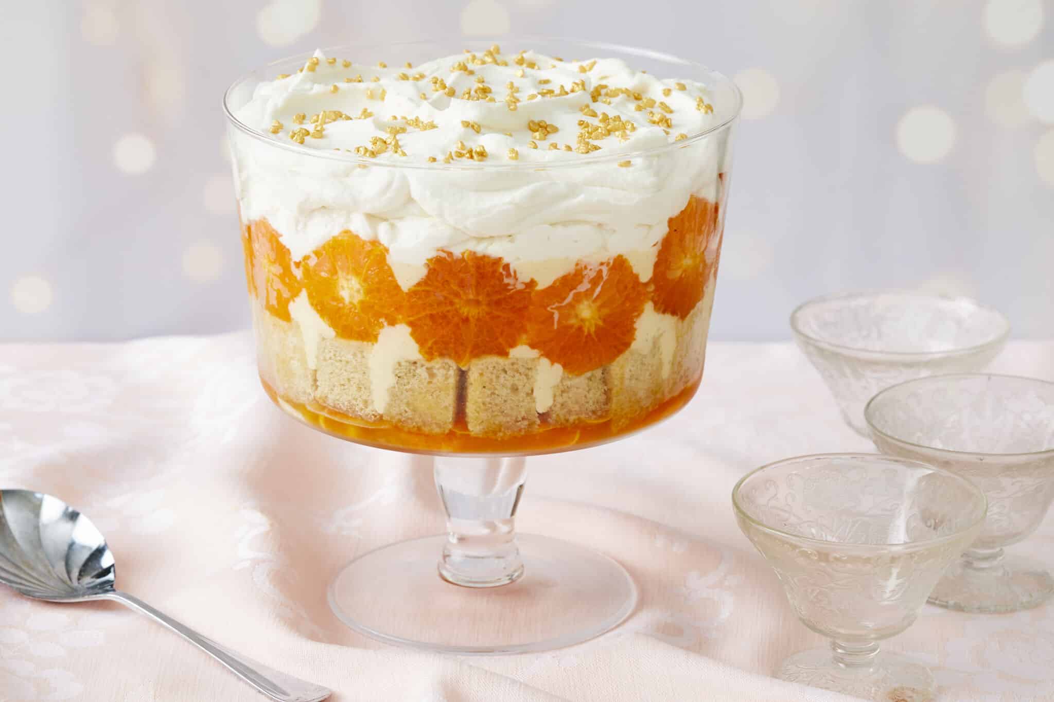 A stunning Sweet and Zesty Clementine Trifle with Boozy Amaretto consists of layers of clementine syrup, cubed sponge cake, caramelized clementines, crème anglaise, lemon curd, and Amaretto whipped cream. Three glass dessert cups and one serving spoon are on the side.