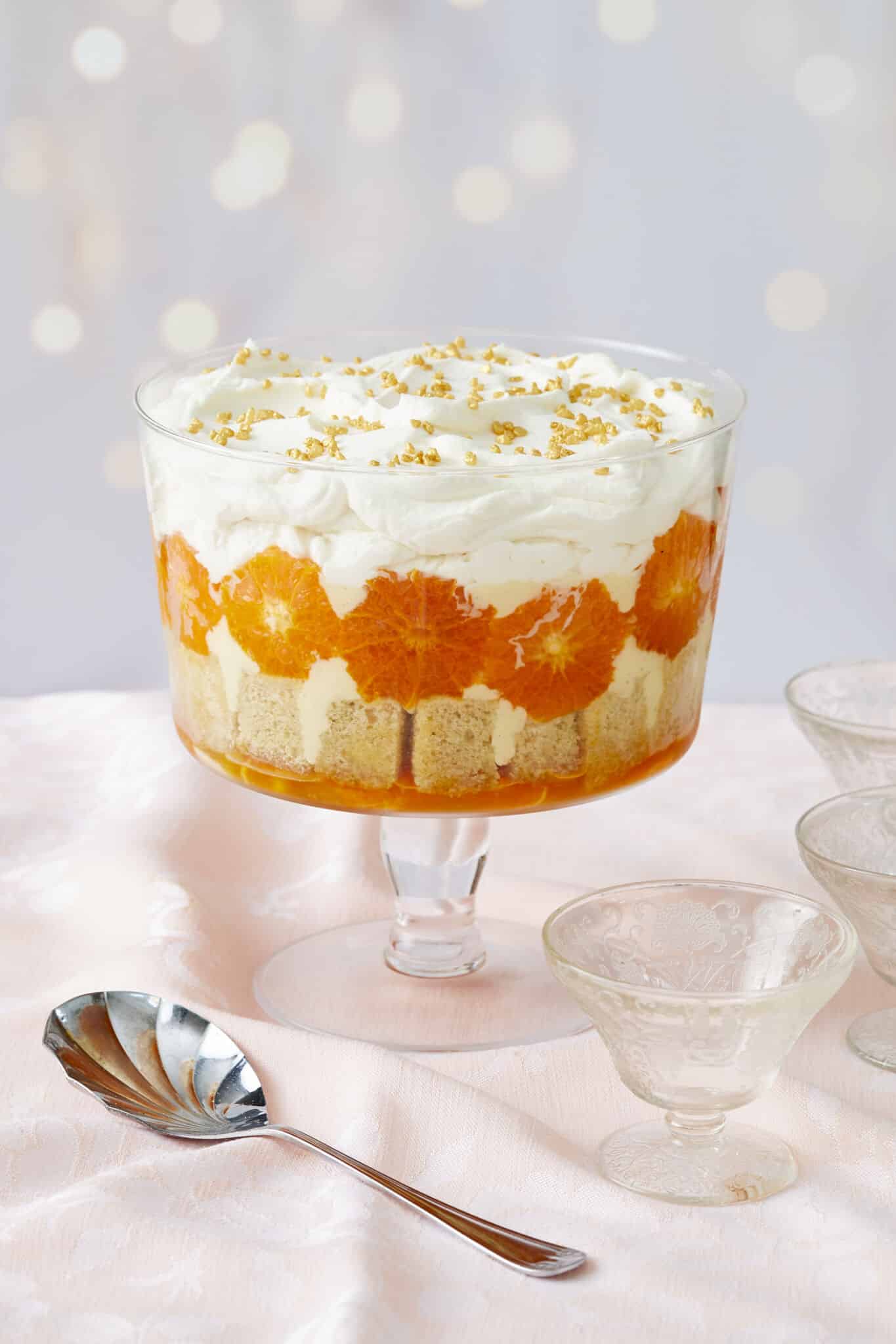 A stunning Sweet and Zesty Clementine Trifle with Boozy Amaretto consists of layers of clementine syrup, cubed sponge cake, caramelized clementines, crème anglaise, lemon curd, and Amaretto whipped cream. Three glass dessert cups and one serving spoon are on the side. 