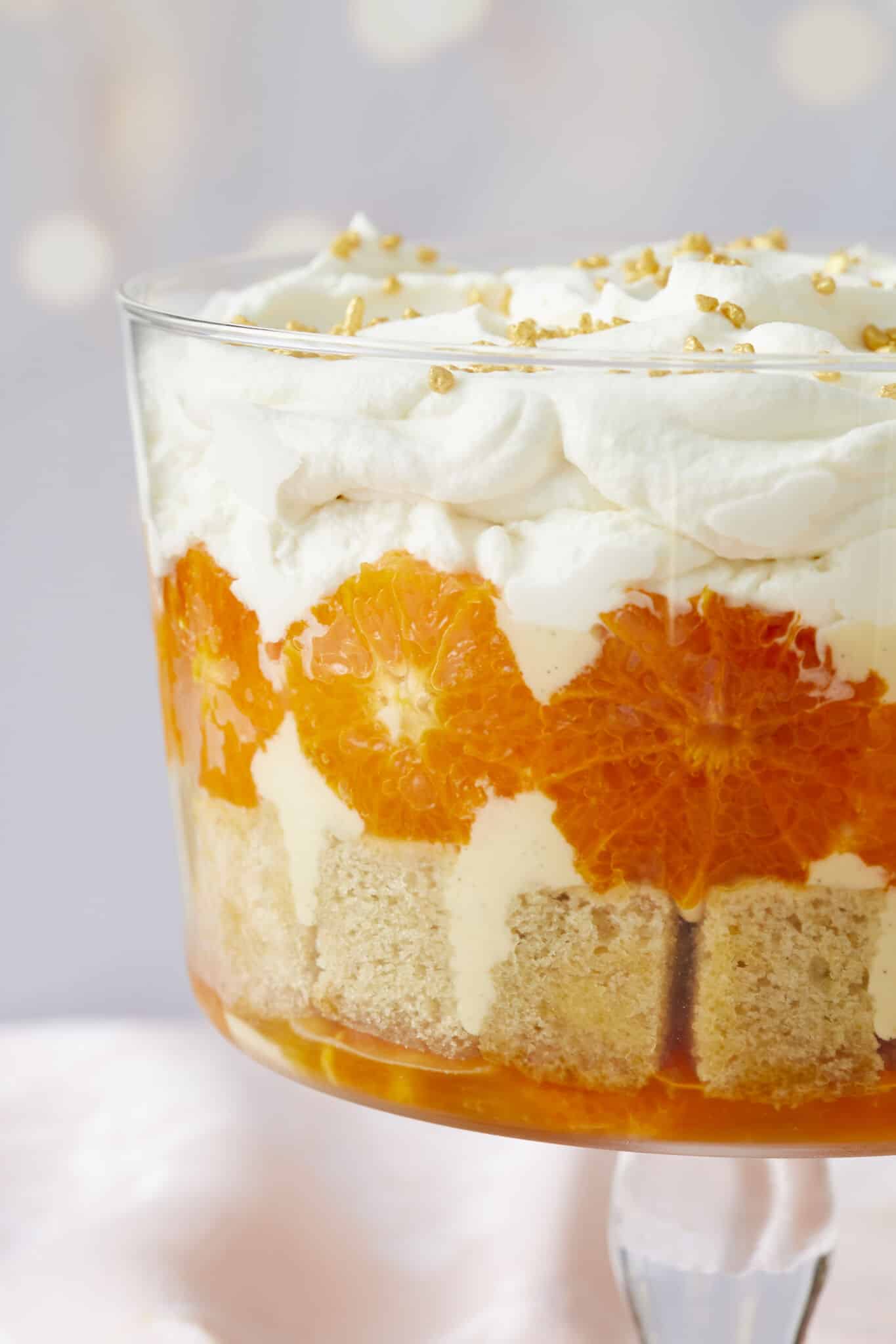 A close-up shot at the stunning Sweet and Zesty Clementine Trifle with Boozy Amaretto shows layers of clementine syrup, cubed sponge cake, caramelized clementines, crème anglaise, lemon curd, and Amaretto whipped cream. Three glass dessert cups and one serving spoon are on the side. 