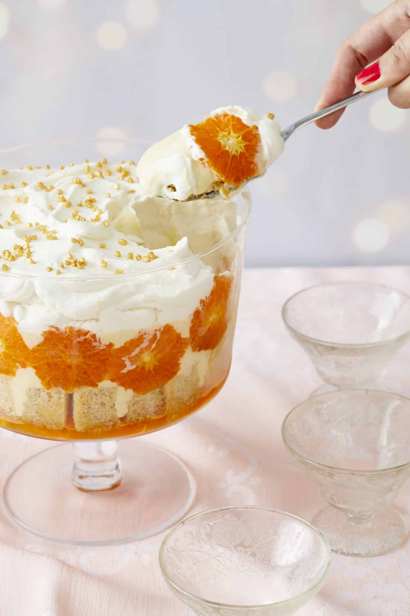 A spoonful of the stunning Sweet and Zesty Clementine Trifle with Boozy Amaretto is being lifted from the bowl. It shows layers of clementine syrup, cubed sponge cake, caramelized clementines, crème anglaise, lemon curd, and Amaretto whipped cream. Three glass dessert cups are on the side.