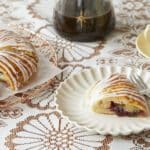 A gorgeous Danish Kringle is golden brown on top gleaming with zig zags of creamy icing. A slice is served on a white dessert plate, showing the swirly and soft inside filled with sweet-tart jam. It's paired with coffee on the side.