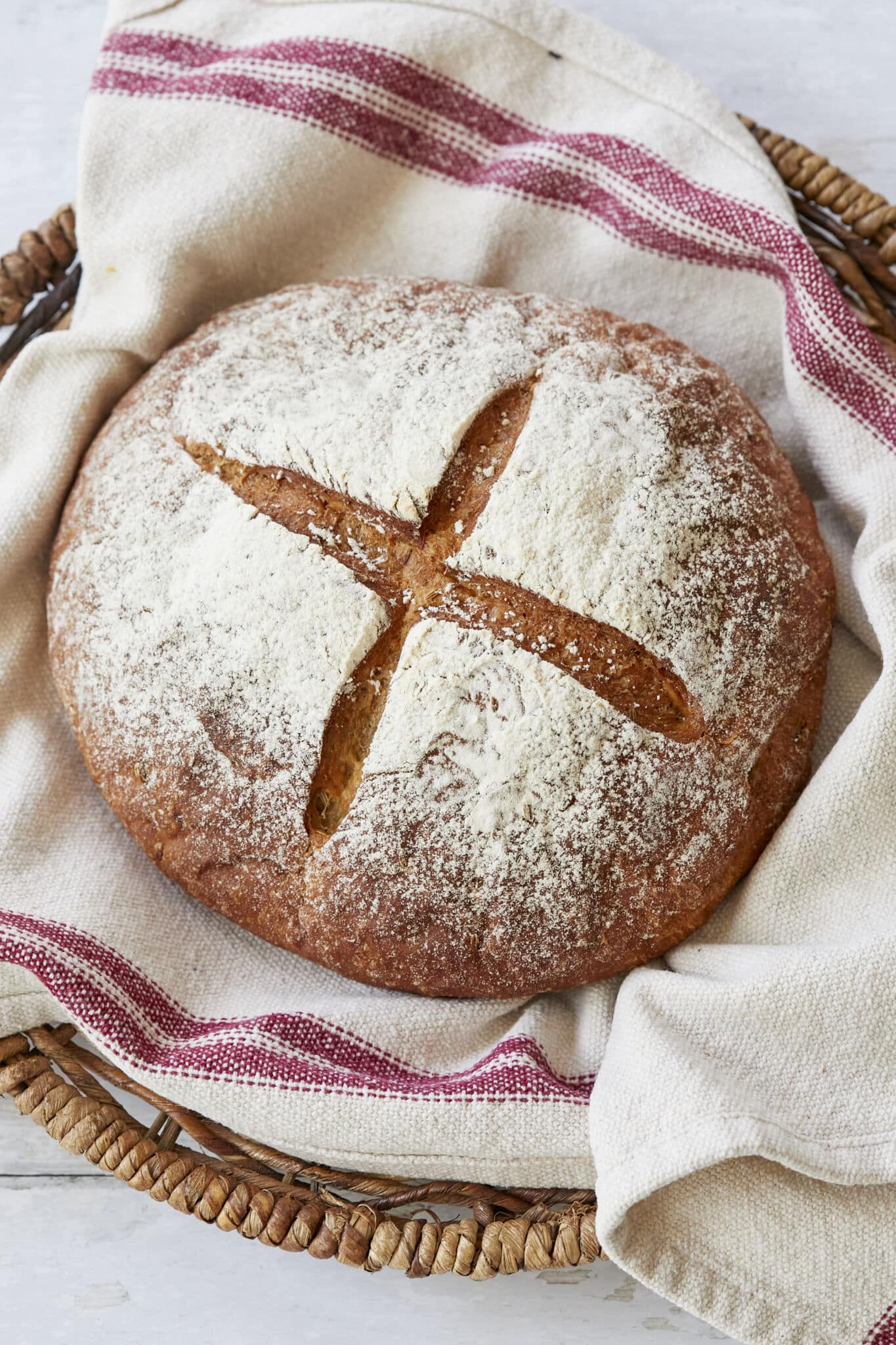 Joululimppu (Finnish Christmas Bread) is a spiced, subtly sweet rye bread and is baked in a round shape scored cross on top. It's dusted with flour and cooling in a basket lined with a kitchen towel. It's in dark brown reddish color. 
