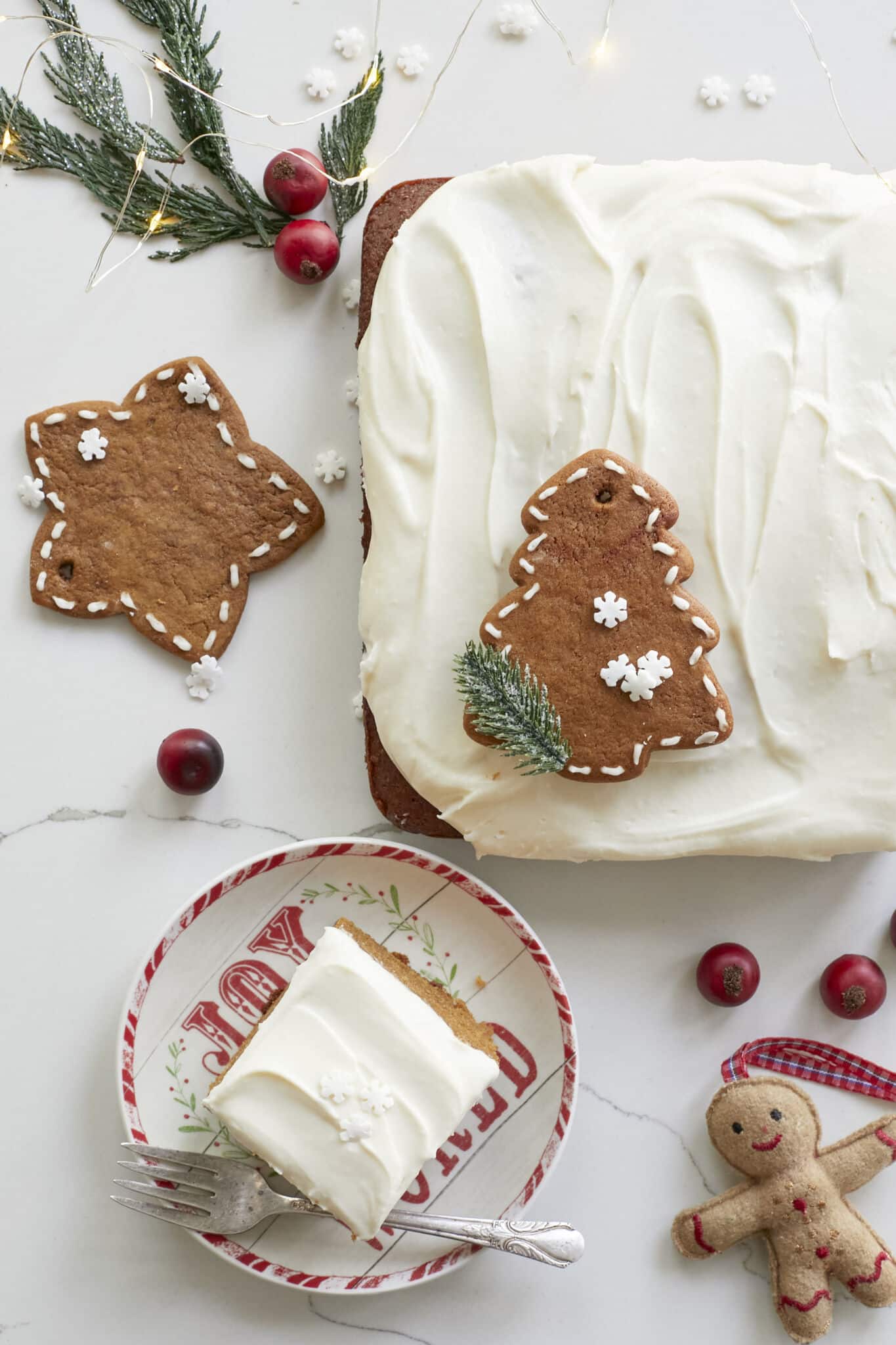 A Perfectly-Spiced Gingerbread Sheet Cake looks moist and soft, topped with velvety cream cheese frosting. A star-shape gingerbread cookie is next to the cake and a Christmas tree shape gingerbread cookie is on the cake for decoration. A generous square of it is served on a Christmas-themed dessert plate. 