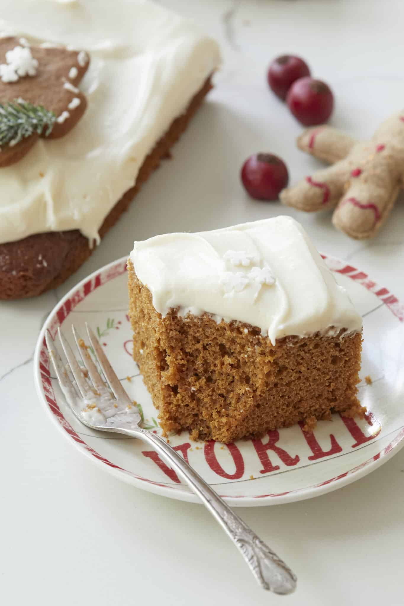 A generous square of Gingerbread Sheet Cake is topped with white frosting and served on a Christmas-themed dessert plate. A bite has been taken. 