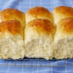 A close shot at Mashed Potato Dinner rolls which are cooling or a wire rack shows their soft and bubbly crumb and golden brown top. The rolls are served with butter on a small plate.
