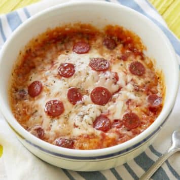 A 2-Minute Microwave Pizza Bowl is cooked to perfection in a big white ceramic bowl with a blue strip near the rim. The pizza is loaded with red sauce, gooey mozzarella cheese, crispy mini pepperoni, and topped with dried herb seasoning.