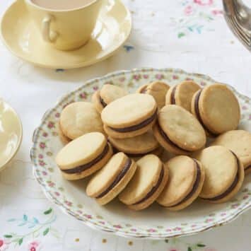 Sandwich cookies Orange Shortbread with Chocolate Orange Truffle Filling are perfectly round. They are served in a large floral platter with two cups of tea.