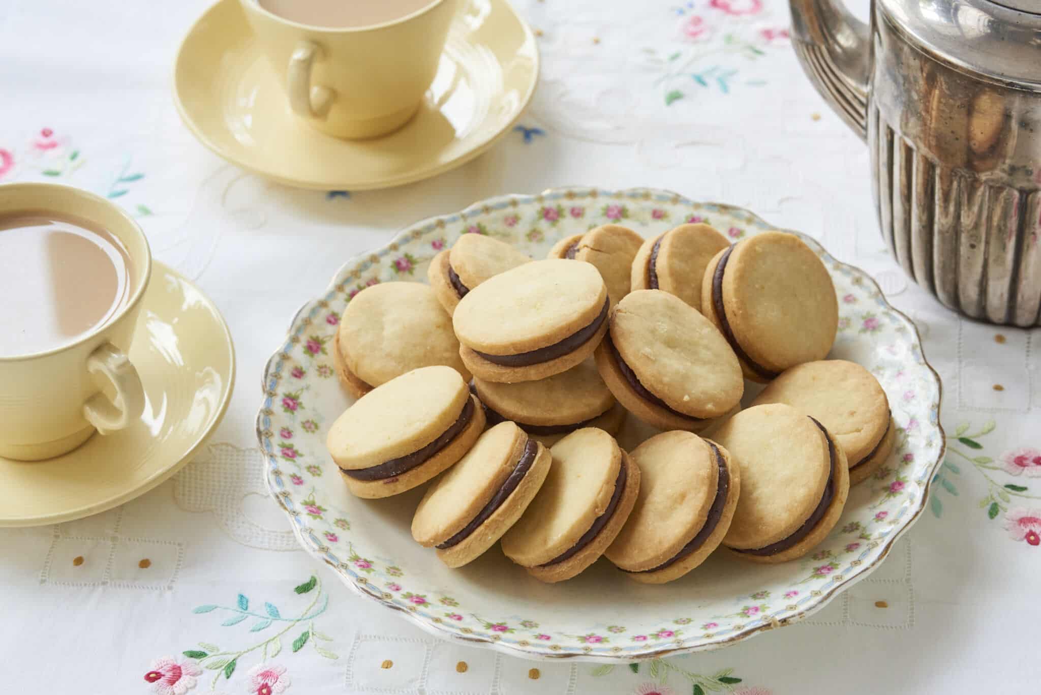 Sandwich cookies Orange Shortbread with Chocolate Orange Truffle Filling are perfectly round. They are served in a large floral platter with two cups of tea.