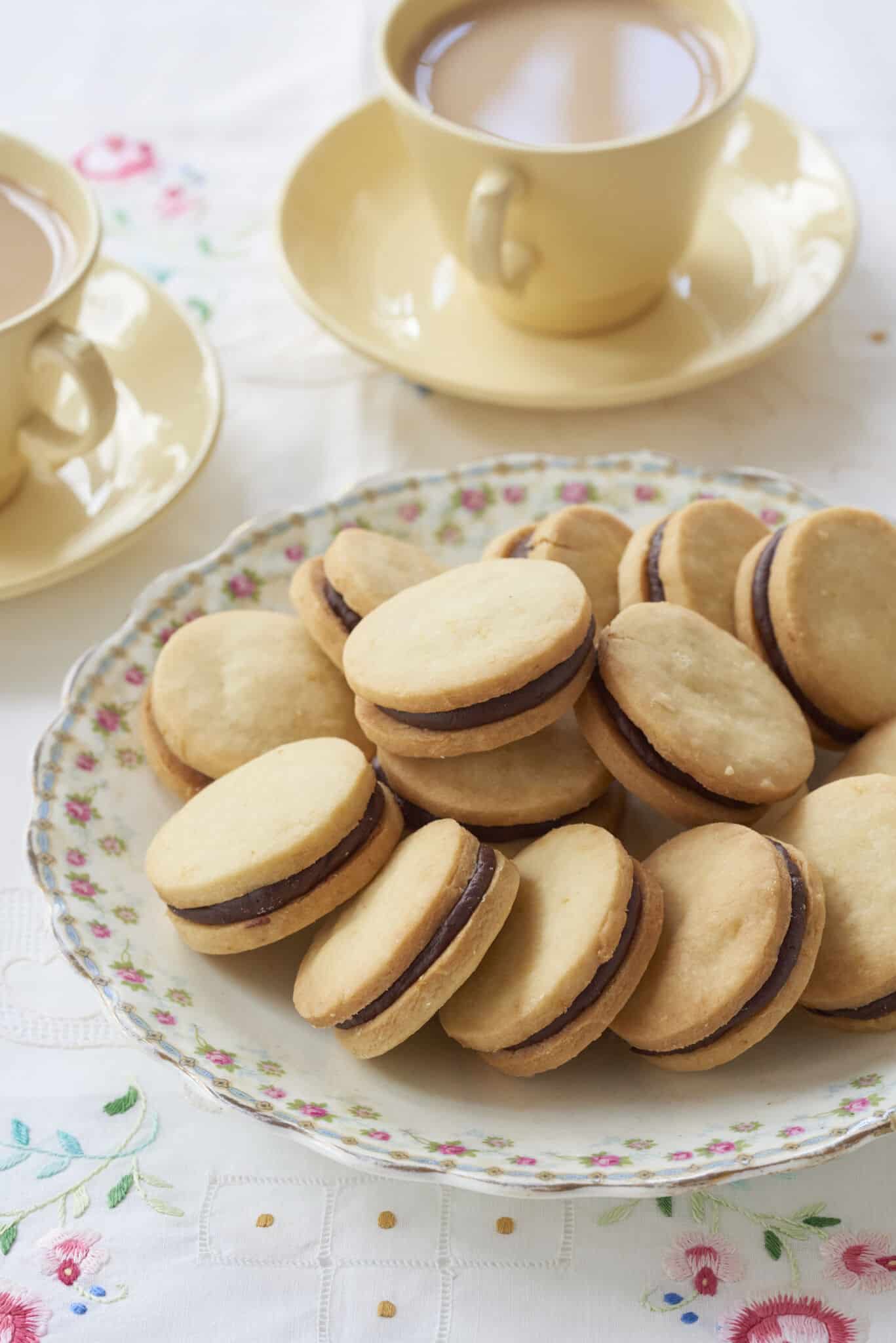 Sandwich cookies Orange Shortbread with Chocolate Orange Truffle Filling are perfectly round. They are served in a large floral platter with two cups of tea. 