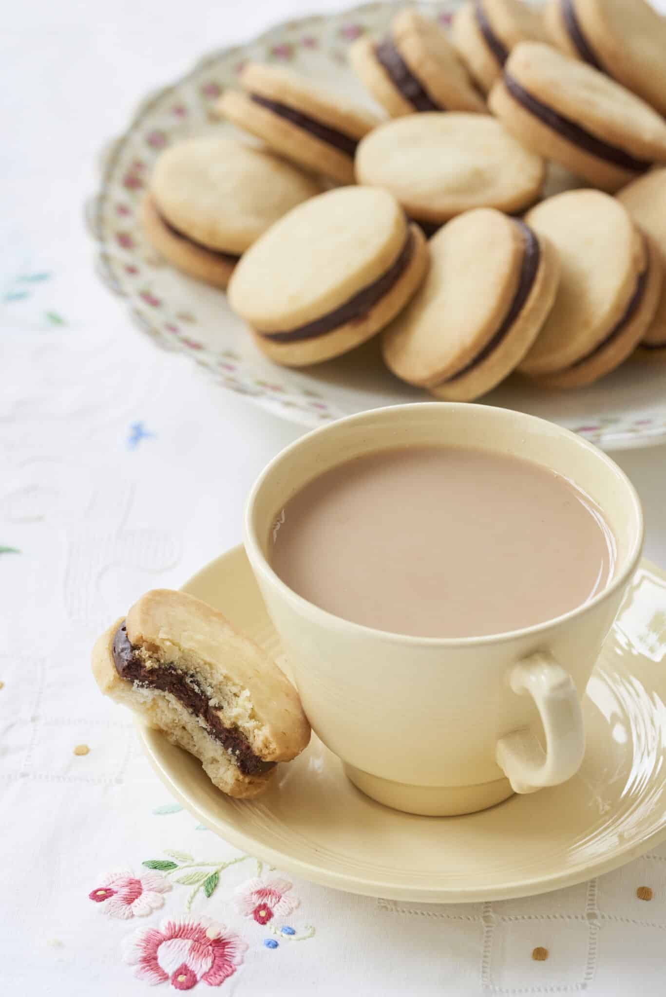Sandwich cookies Orange Shortbread with Chocolate Orange Truffle Filling are perfectly round. One shortbread which has had one bite taken is served with a cup of tea on the saucer. The rest of the shortbread are in a large floral platter on the side. 