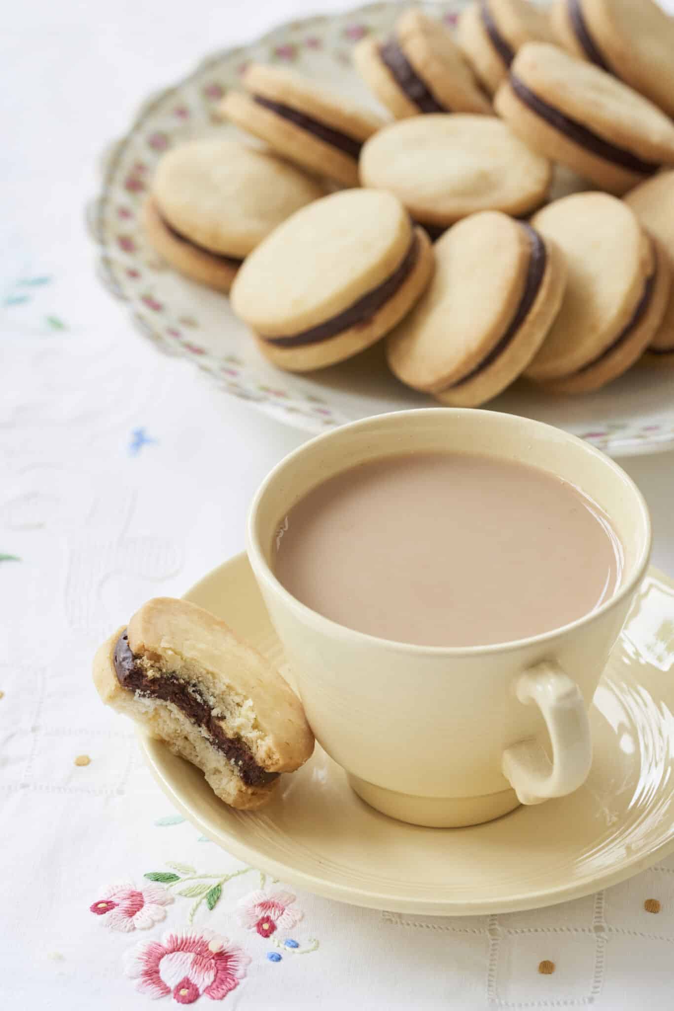 Sandwich cookies Orange Shortbread with Chocolate Orange Truffle Filling are perfectly round. One shortbread which has had one bite taken is served with a cup of tea on the saucer. The rest of the shortbread are in a large floral platter on the side.