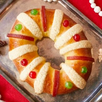 Rosca de Reyes (Three Kings Bread)is baked until golden brown. It's infused with orange zest and decorated with festive candied cherries, strips of quince paste, and sugar paste. A baby Jesus figurine is inserted into the bread after baking,