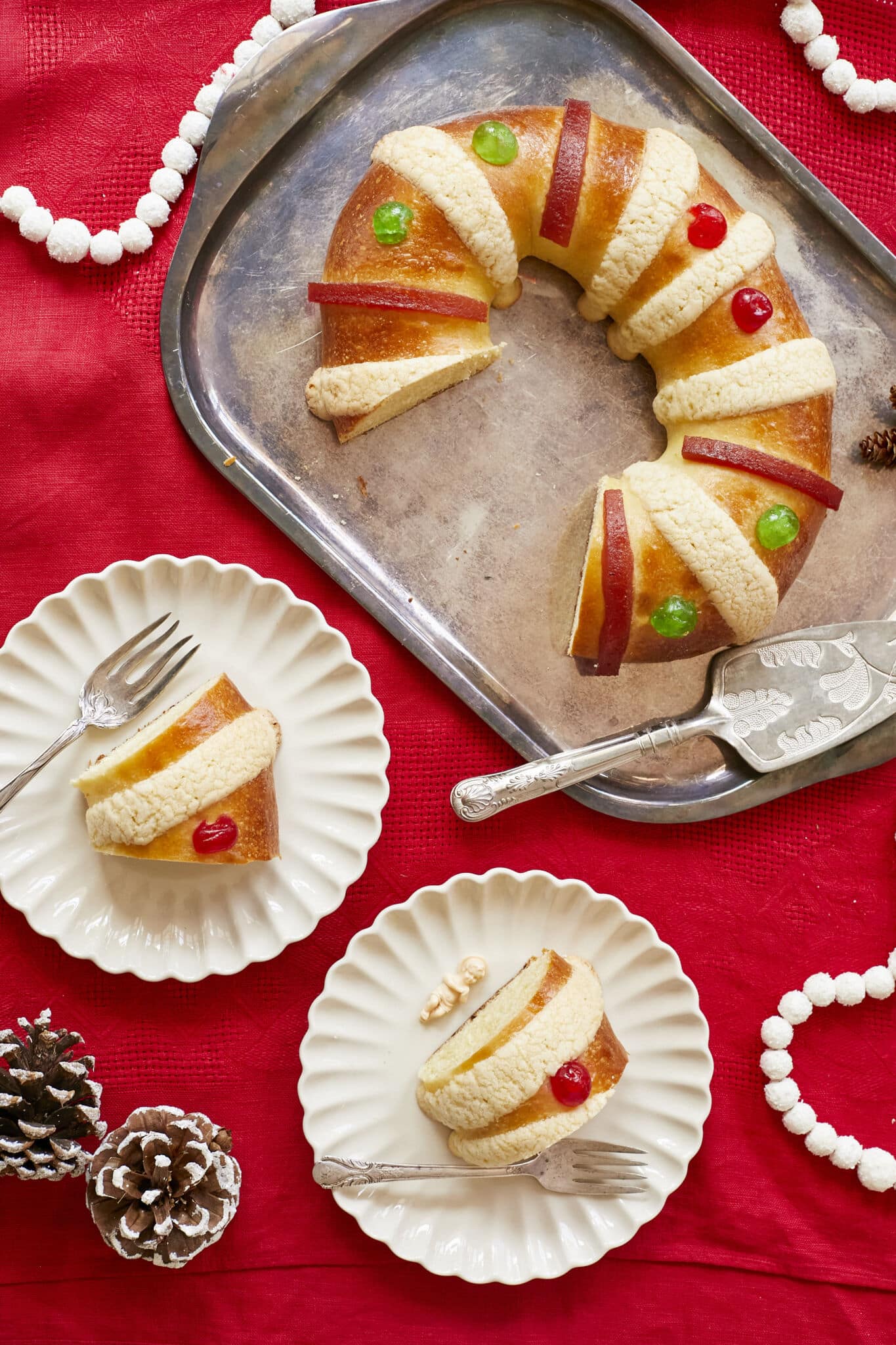 Rosca de Reyes (Three Kings Bread)is baked until golden brown. It's infused with orange zest and decorated with festive candied cherries, strips of quince paste, and sugar paste. A baby Jesus figurine is inserted into the bread after baking, Two slices are cut from the whole bread and served in dessert plates.