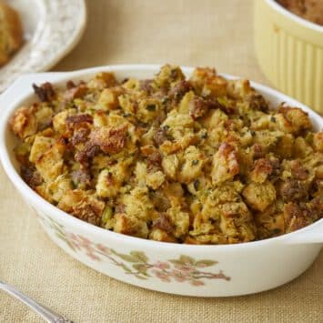 Classic Sage Sausage Stuffing is baked in a white milk galss baking dish. The dressing is alsos erved on a big golden-rim platter and a yellow baking dish. Toasted white bread is tossed with savory sausage, earthy sage, and aromatic onion and celery, mixed with in eggs and broth to a fluffy, moist casserole with tempting craggy browned bits!