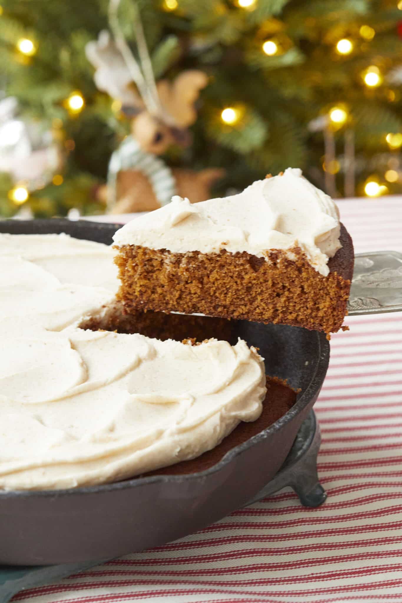 A slice of rich, moist Gingerbread Cake is being lifted from the cast iron skillet, crowned with velvety Brown Butter Frosting. A Christmas Tree with lights on is in the background. 
