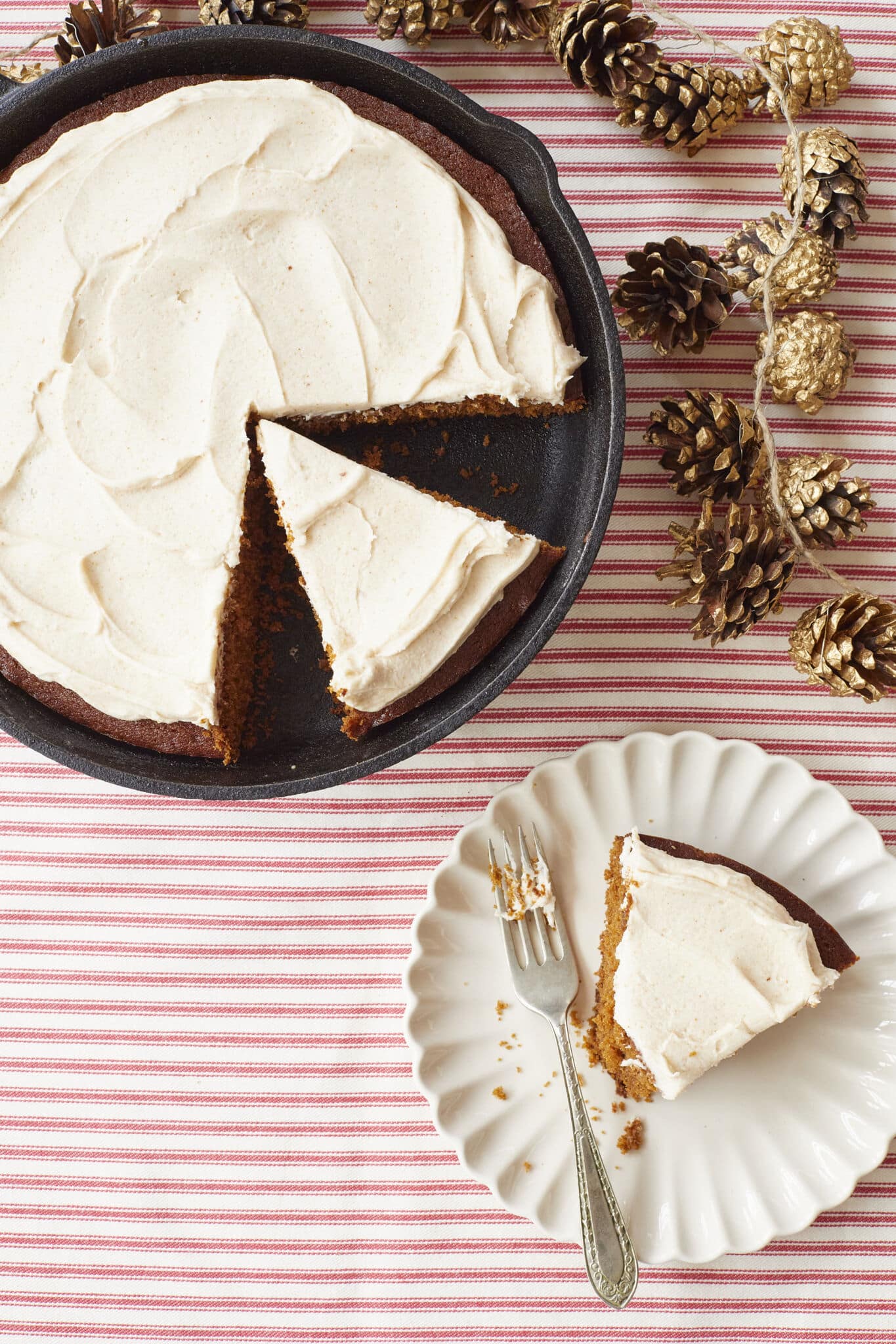 Rich, moist Gingerbread Cake is baked in a cast iron skillet, crowned with velvety Brown Butter Frosting. A slice is served on a dessert plate. Some dried pine cones are decorated on the side.