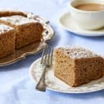 The Traditional Irish Spice Cake is cut into generous squares and served on a silver platter. They have delectably moist, fine crumb and are dusted with powdered sugar. One slice is served on a floral dessert plate, paired with a cup of tea on the side.