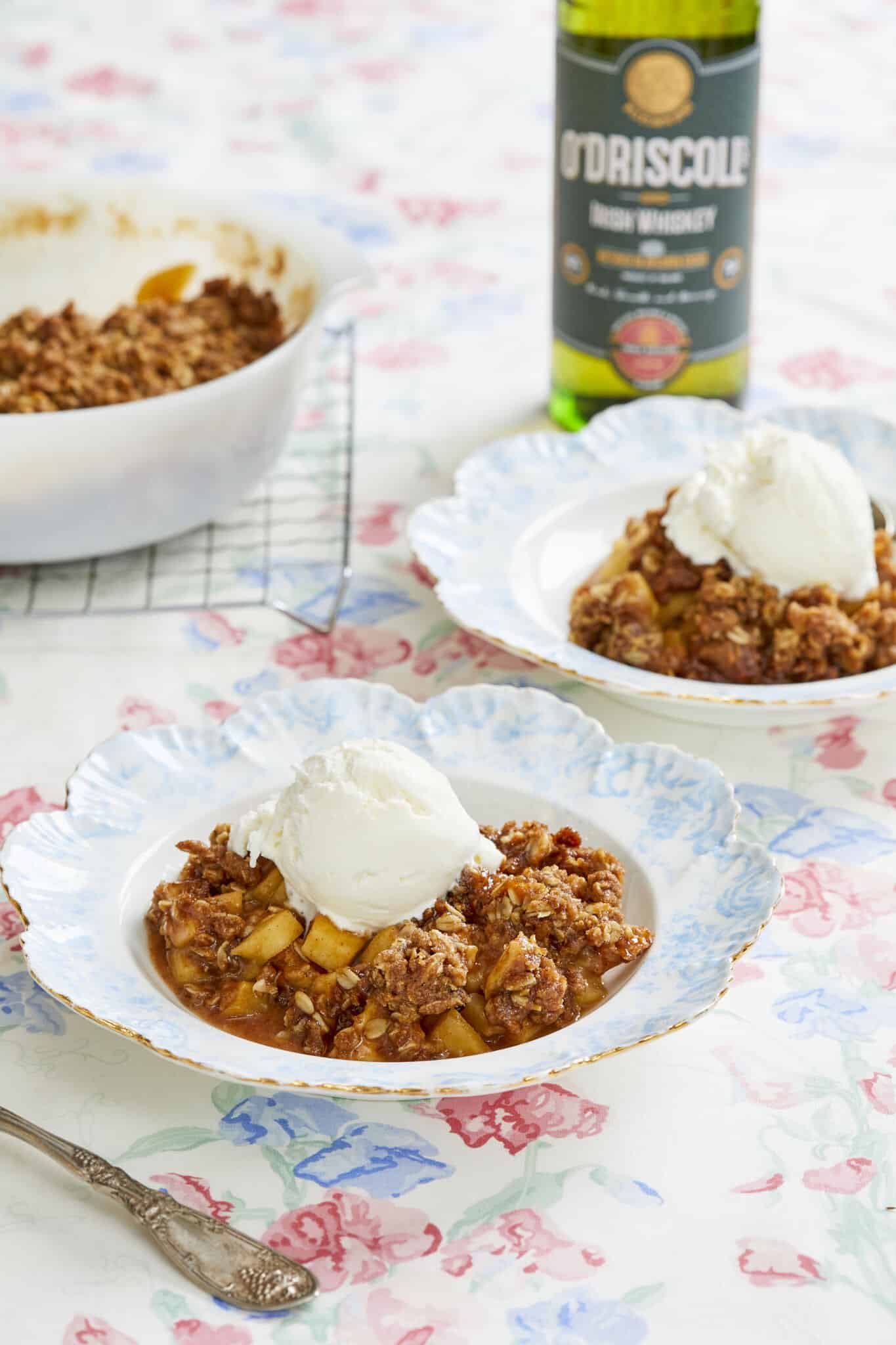 Boozy Whisky Apple Crisp is baked in a milkgalss Pyrex baking dish with sweet, buttery baked apples covered with a cinnamon oatmeal-cinnamon crispy top. Two servings are ready in dessert plates with ice cream on top.