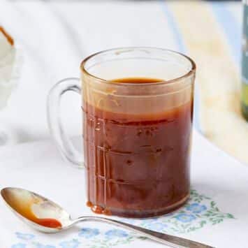 Irish Whiskey Caramel Sauce is in a glass measuring jug with a spoon next to it. It's in deep amber color and very silky smooth consistency. A cup of vanilla ice cream crowned with this caramel sauce is served on the side and the rest of the whiskey is in its green O'Driscolls bottle.