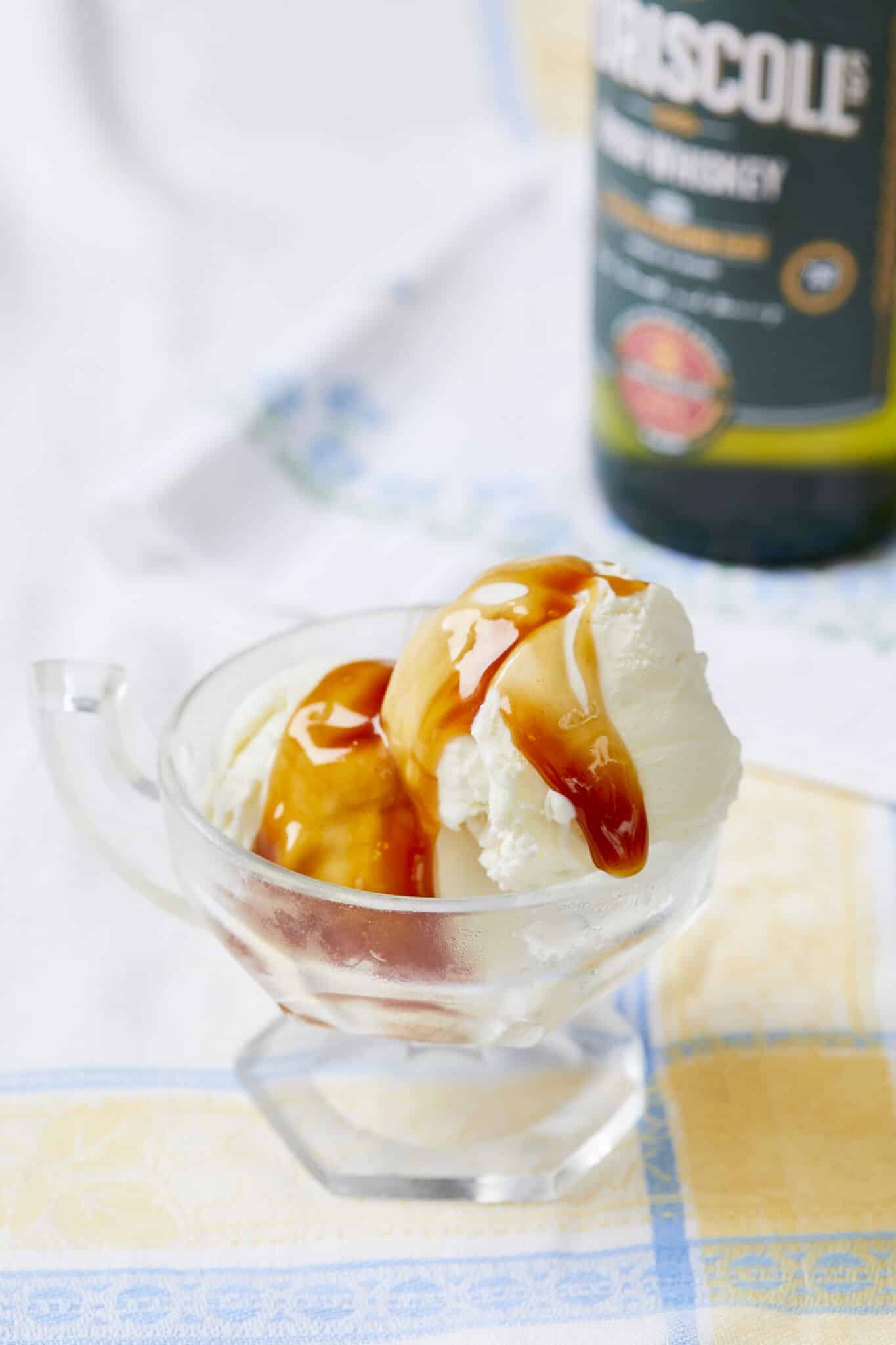 Deep-amber, silky smooth Irish Whiskey Caramel sauce is drizzled on two generous scoops of vanilla ice cream in a glass ice cream cup. A bottle of O'Dricsolls Irish whiskey is on the side. 