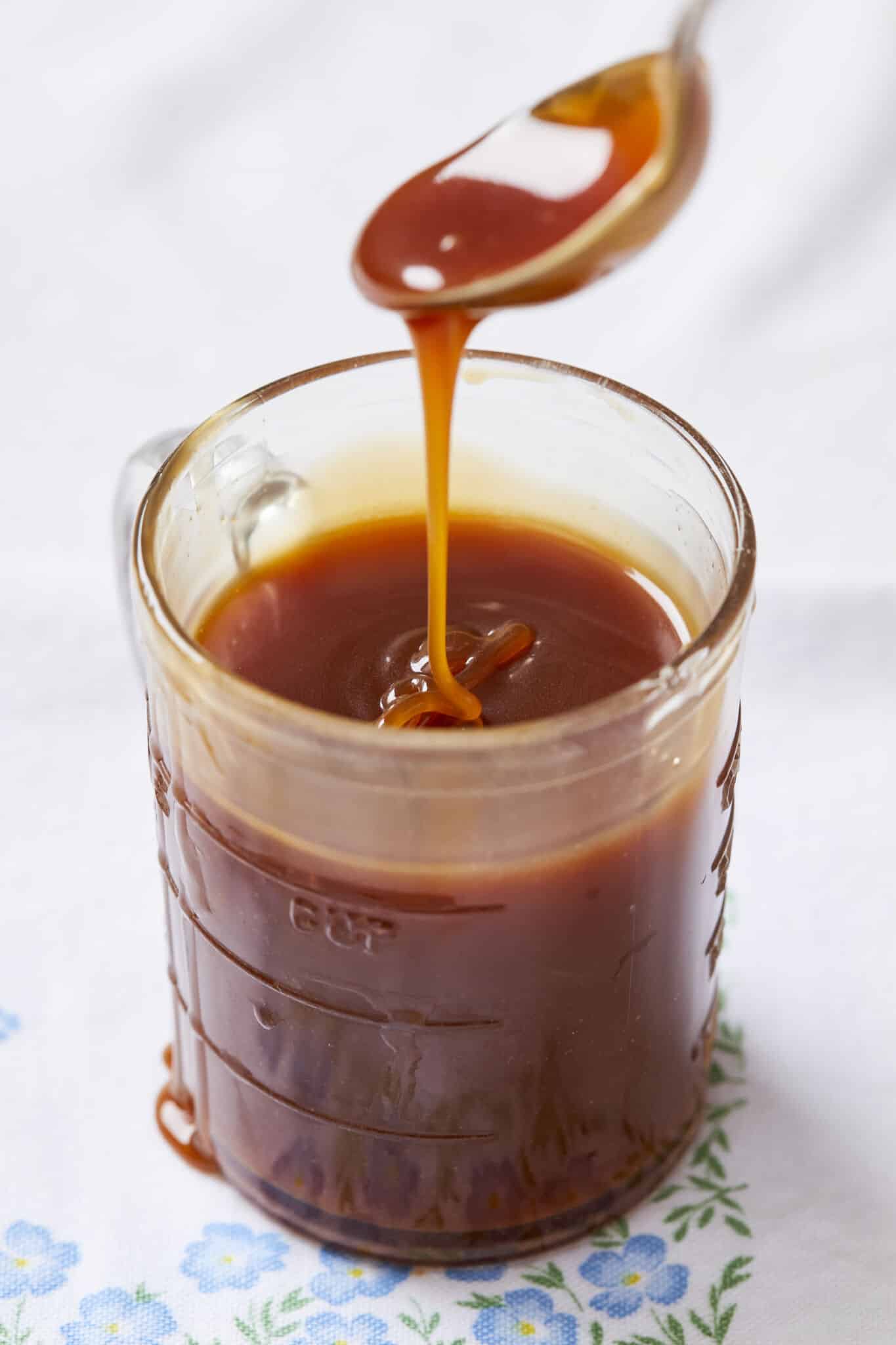 Irish Whiskey Caramel Sauce is in a glass measuring jug. It's in deep amber color and very silky smooth consistency. The whiskey caramel sauce is dripping from a spoon above the jug back in.