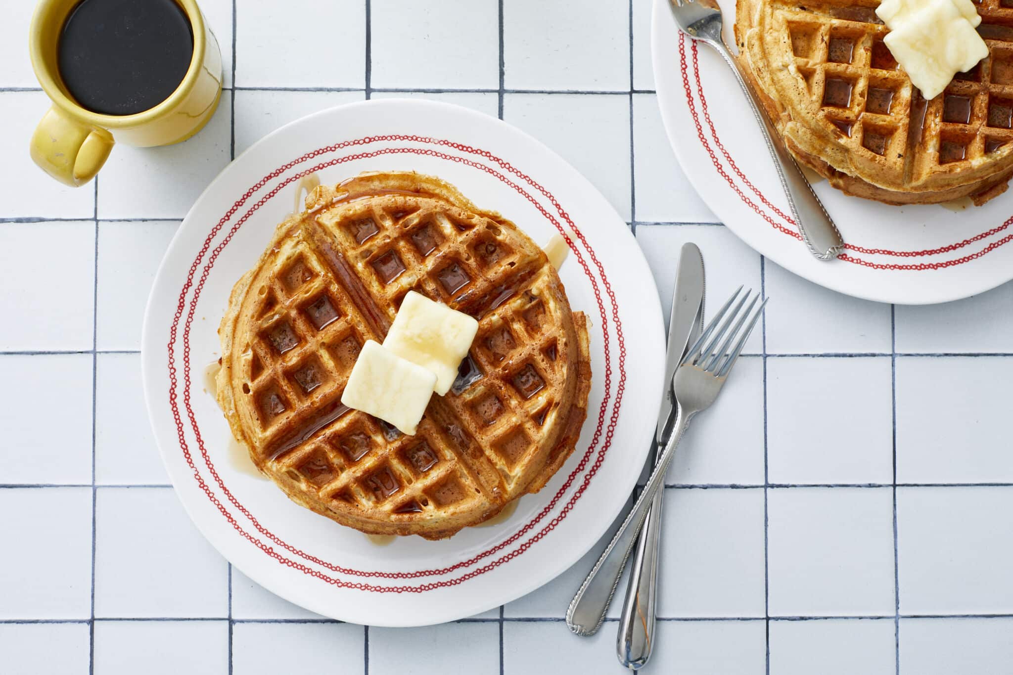 An overhead shot at big golden, fluffy and crispy Whole Wheat Waffles with Wheat Germ served on plates with butter and maple syrup. A cup of coffee is on the side.