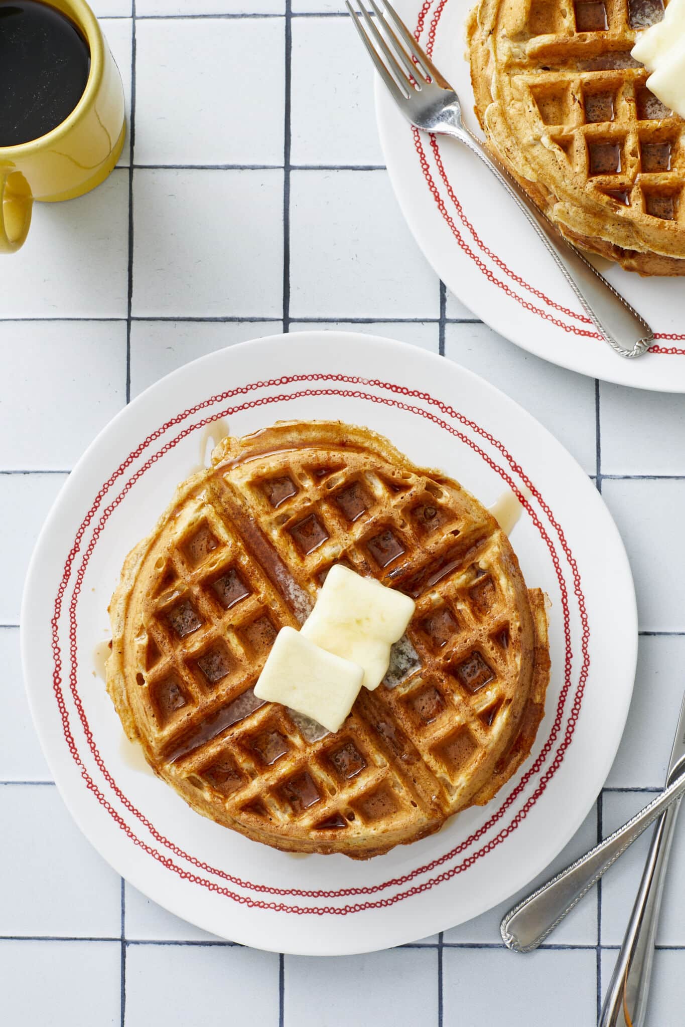 An overhead shot at big golden, fluffy and crispy Whole Wheat Waffles with Wheat Germ served on plates with butter and maple syrup. A cup of coffee is on the side. 