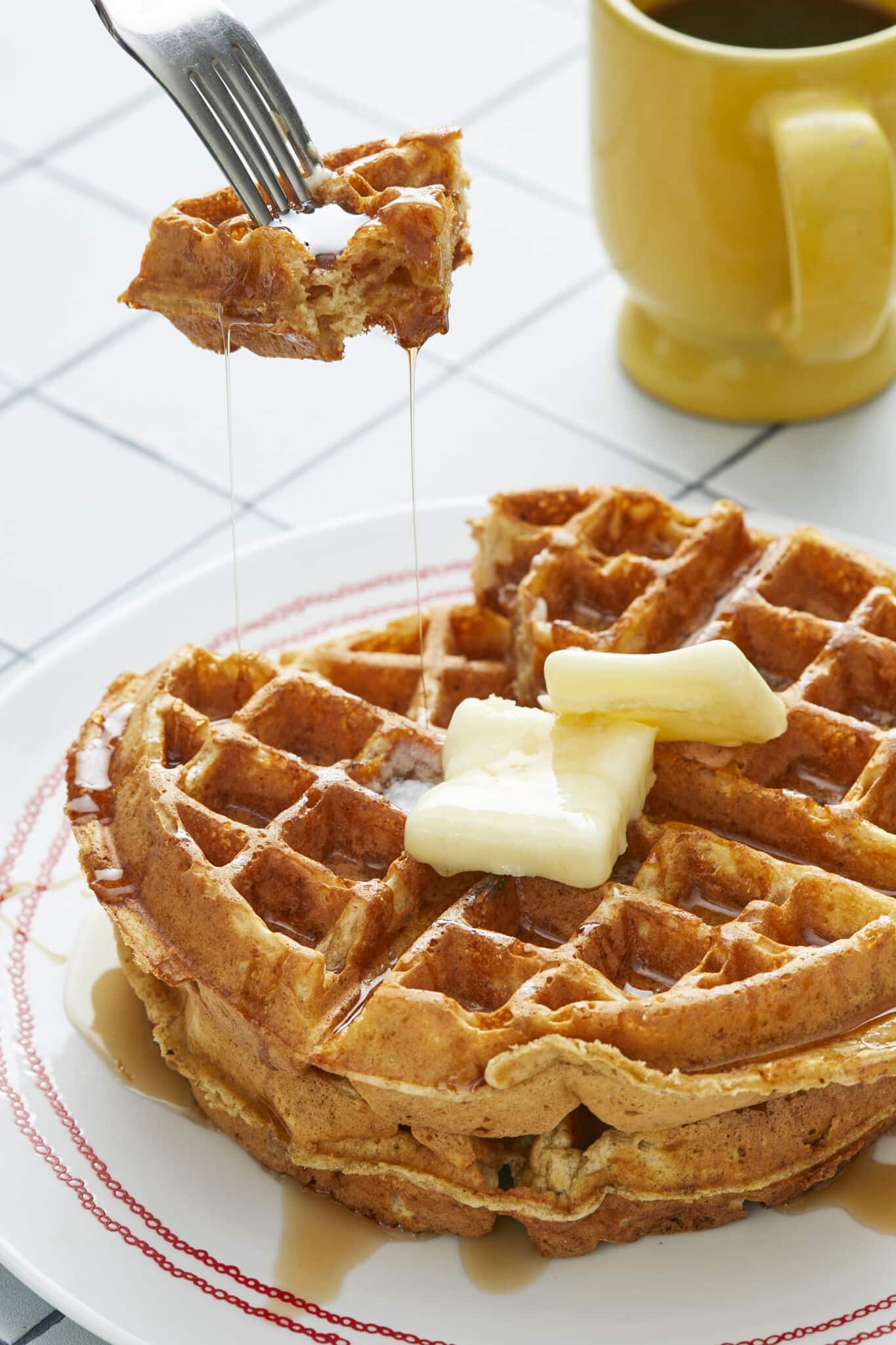Two big golden, fluffy and crispy Whole Wheat Waffles with Wheat Germ are served on a plate with butter. A bite has been cut and is being lifted from the waffles with maple syrup dripping off. 