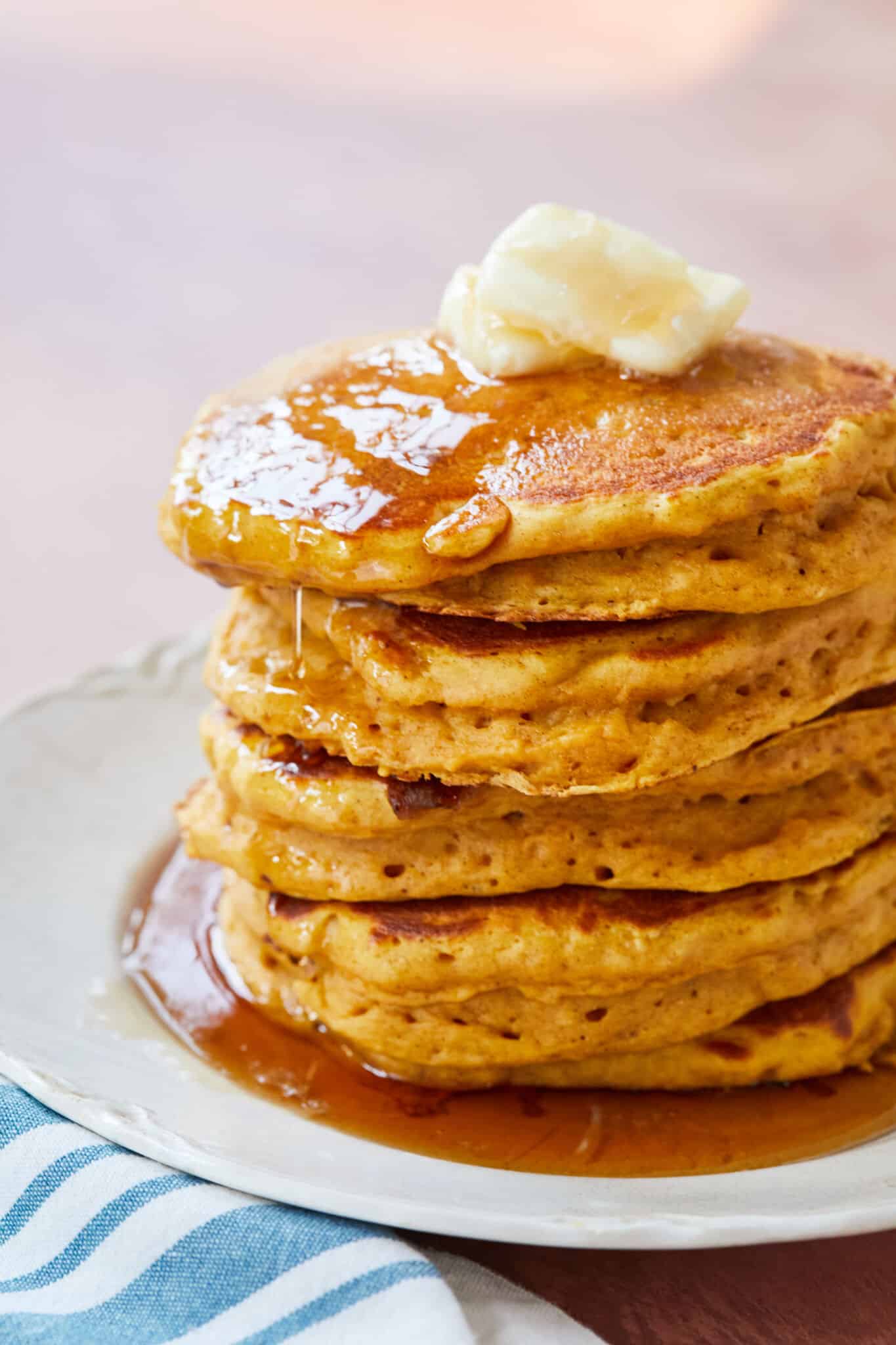 A stack of Pumpkin Buttermilk Pancakes are cooked to perfection with evenly-distributed bubbles, looking very soft, fluffy, and moist. They're seven with butter and generous amount of maple syrup as a scrumptious holiday breakfast or any day breakfast! 