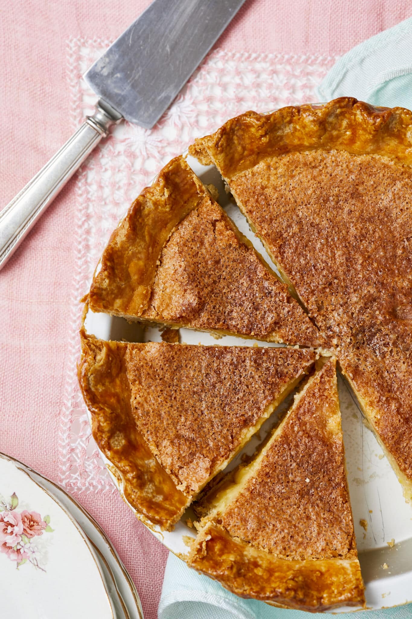 A Classic Chess Pie is baked to perfection with golden brown color all around the edge and the surface of the filling. The crust is very flaky and the caramelized topping is crispy. Half of the pie is cut into four slices and one slice has been removed from the pie dish. 