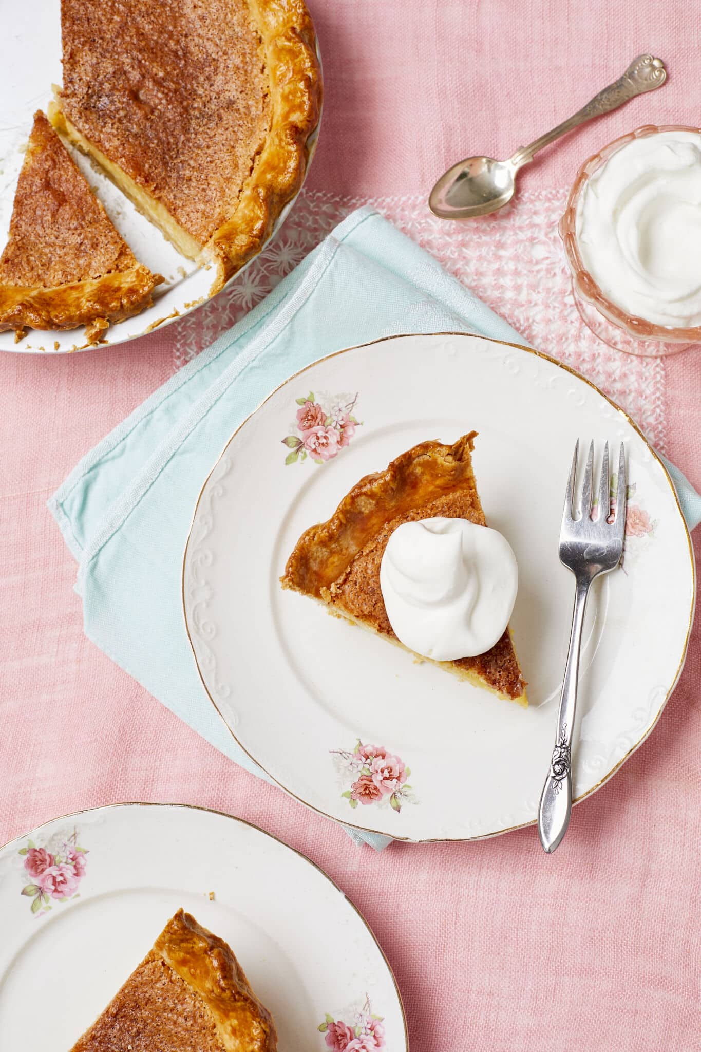 An overhead shot of the Classic Chess Pie. 
Two slices of the Classic Chess Pie are served on golden-rim dessert plates. The slice has flaky crust and crispy caramelized topping crowned with a big dollop of whipped cream. 

The rest of the pie is in the pie pan on the side. Extra whipped cream is served in a pink great depression glass cup. 