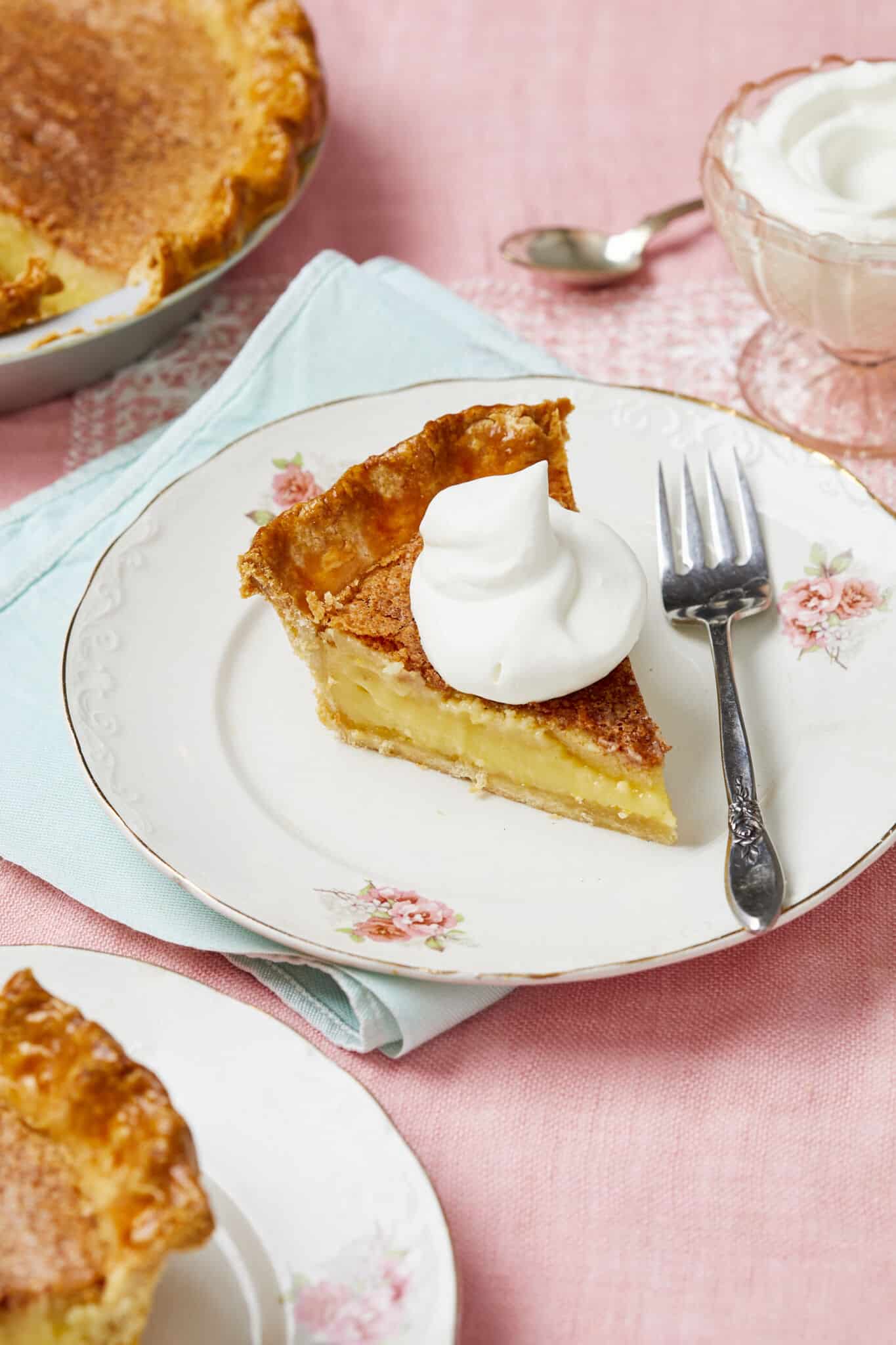 Two slices of the Classic Chess Pie are served on golden-rim dessert plates. The slice has flaky crust, silky filling, and crispy caramelized topping crowned with a big dollop of whipped cream. 

The rest of the pie is in the pie pan on the side. Extra whipped cream is served in a pink great depression glass cup. 