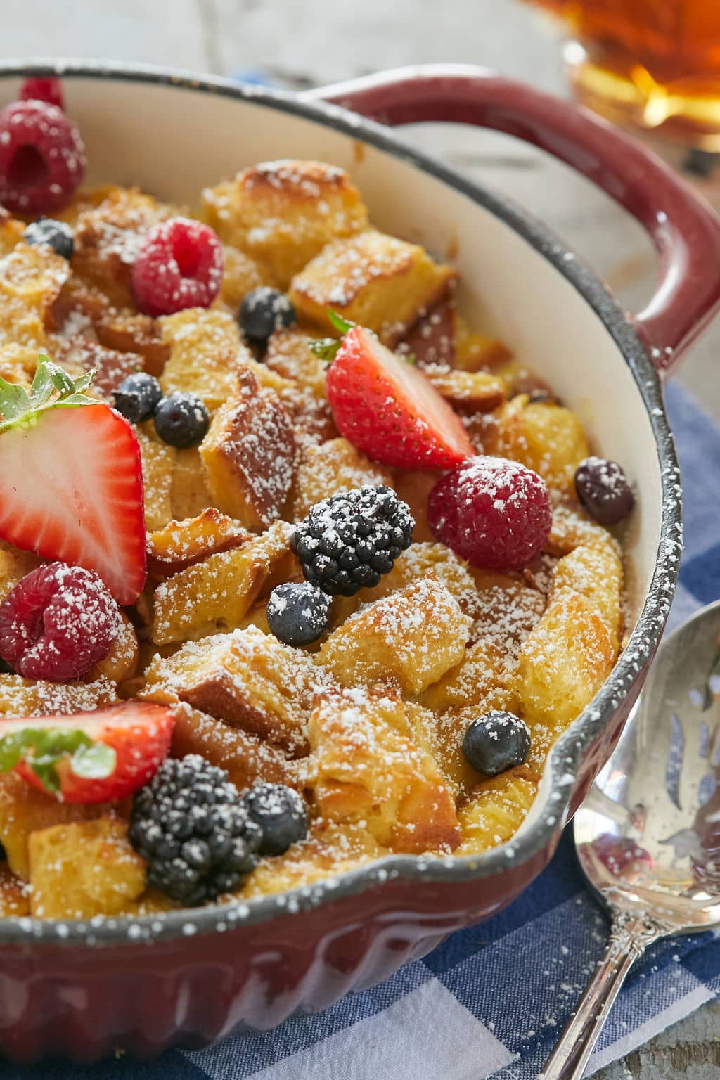 A pan of gorgeous Overnight French Toast has golden crispy toasted bread cube, soft creamy custard, and juicy sweet colorful strawberries, blueberries, and blackberries. It's sprinkled with powdered sugar for extra flavor. 
