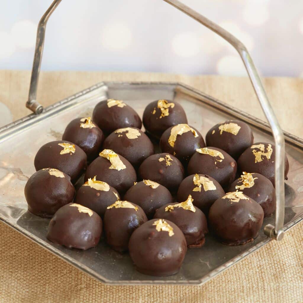 Heavenly Champagne Truffles are placed on a silver platter with a handle. they have bittersweet chocolate shells and are decorated with edible gold leaves.