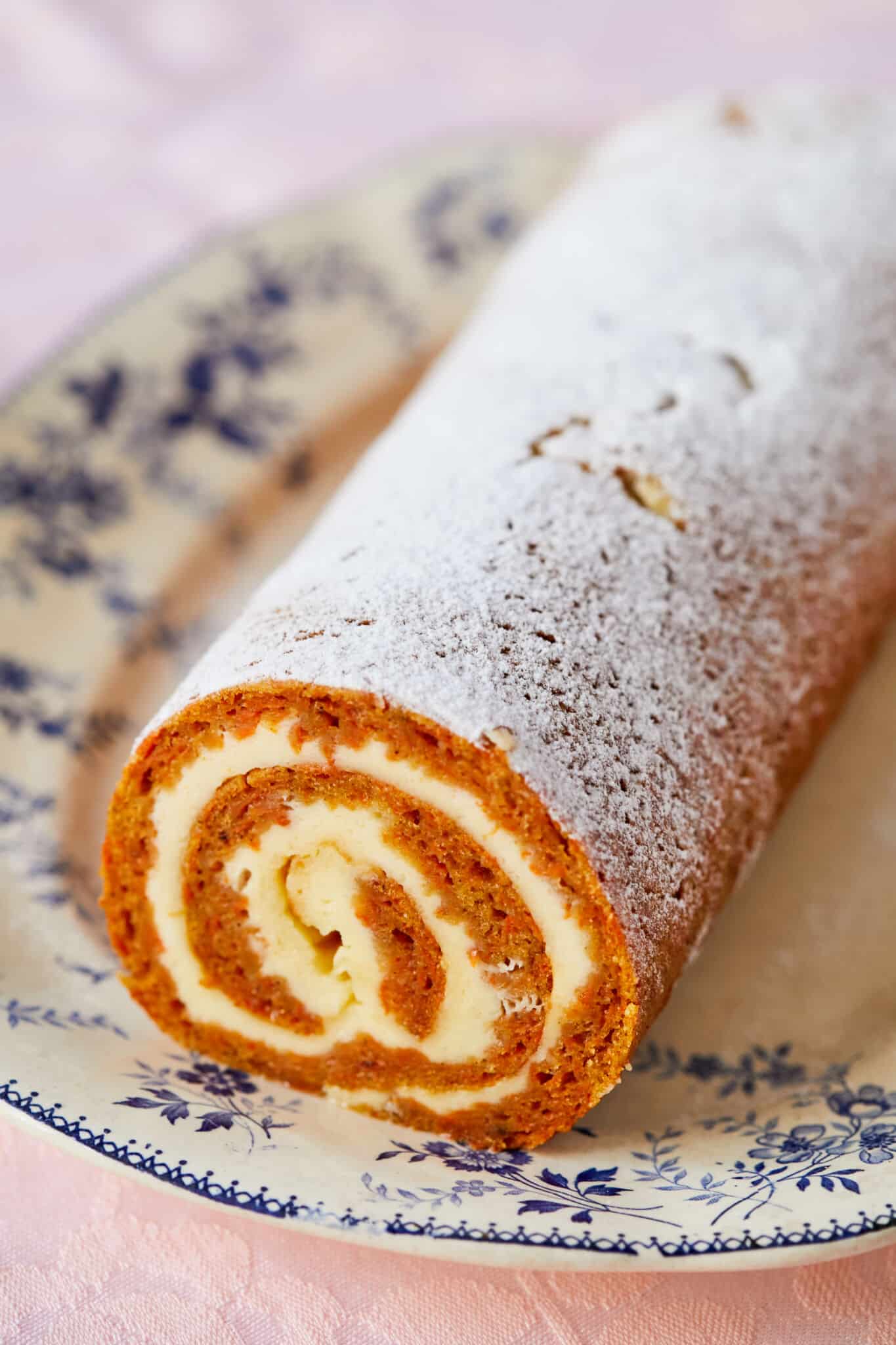 A log of Carrot Cake Cheesecake Roll is served on a floral platter. The cake is moist and soft, stuffed with creamy and tangy cream cheese filling and dusted with powdered sugar. The orange cake and milky white color add a gorgeous color contrast.