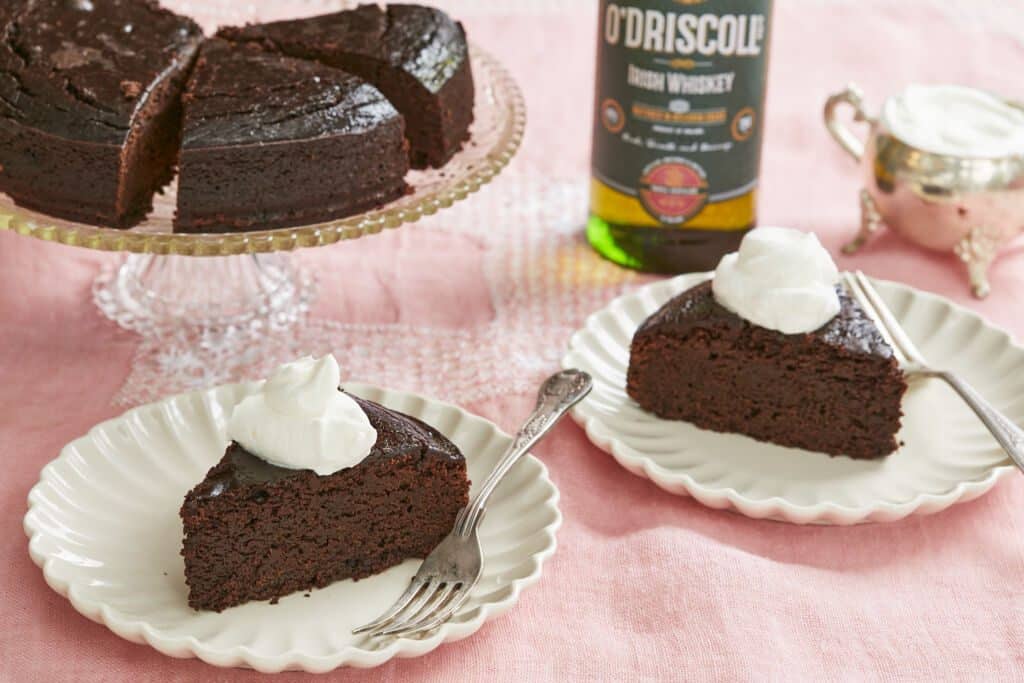 Rich, moist and decadent Irish whiskey Chocolate Cake are sliced and served with whipped cream. A bottle of whiskey and extra whipped cream are on the side.